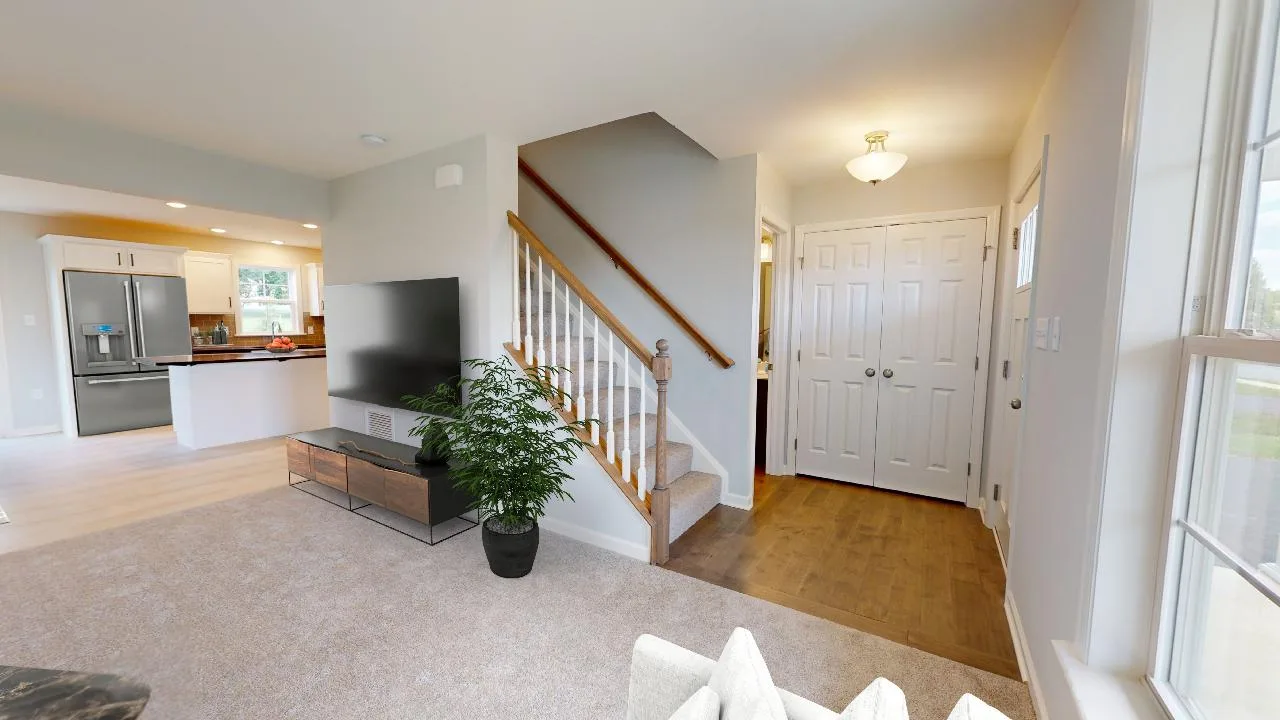 A bright living room with beige carpet, a TV on a wooden stand, a potted plant, a staircase with a wooden railing, and an entryway with double doors and hardwood flooring. The kitchen is visible in the background.