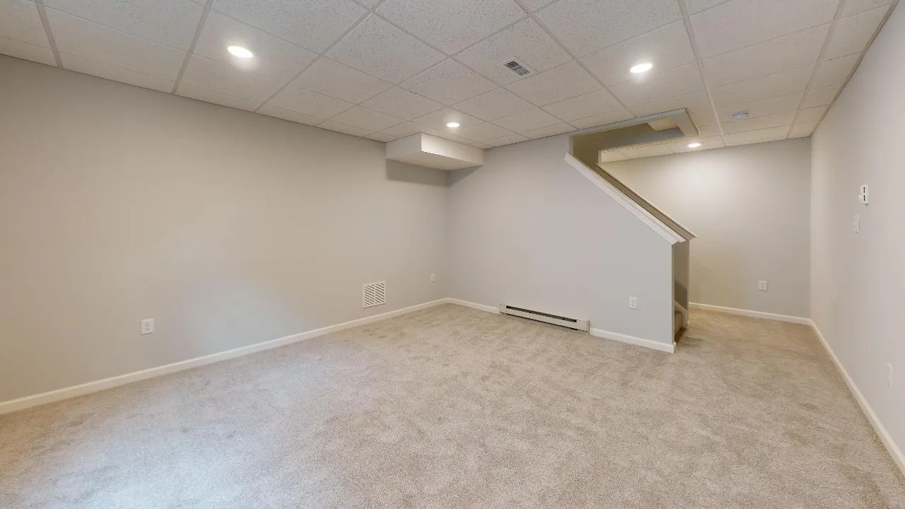 A finished basement with beige carpet, light gray walls, recessed ceiling lights, and a staircase leading up. The space is empty and clean with a modern, neutral design.