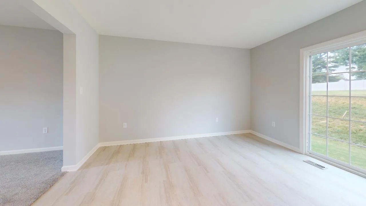 A bright, empty room with light wood floors, pale gray walls, and a large sliding glass door leading to a grassy outdoor area. There is an open entryway connecting to a carpeted room.