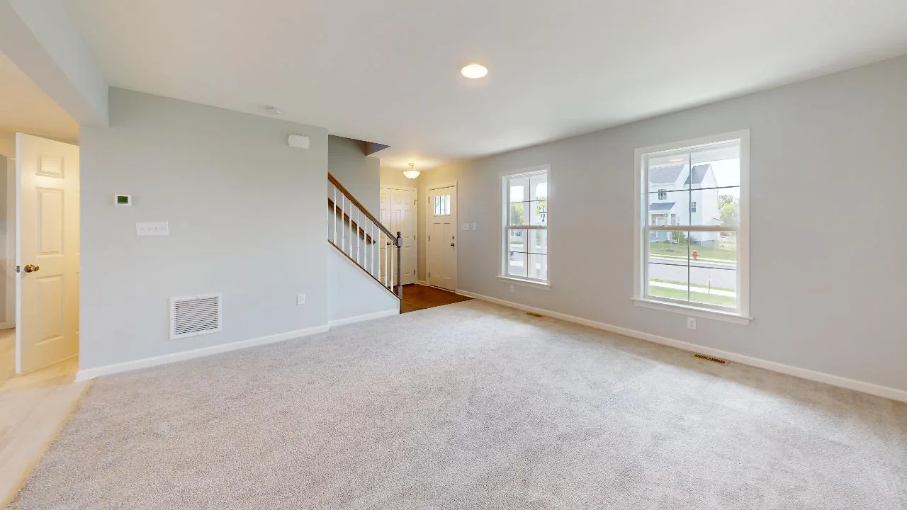 Spacious living room with light gray walls, beige carpet, two large windows, and a staircase leading upstairs. The front door and hallway are visible, allowing natural light to fill the space.
