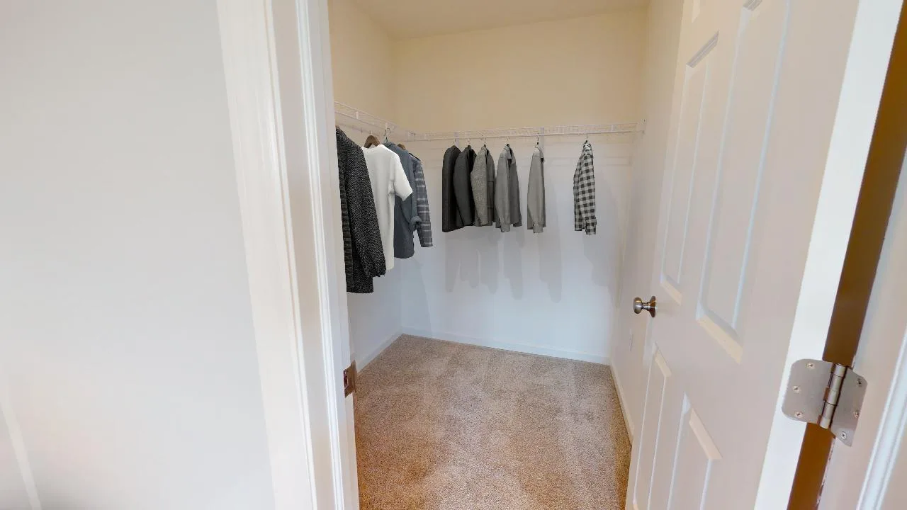 A walk-in closet with beige carpet, white walls, and a wire shelf holding several hanging shirts and jackets, mostly in neutral colors. The closet door is open.