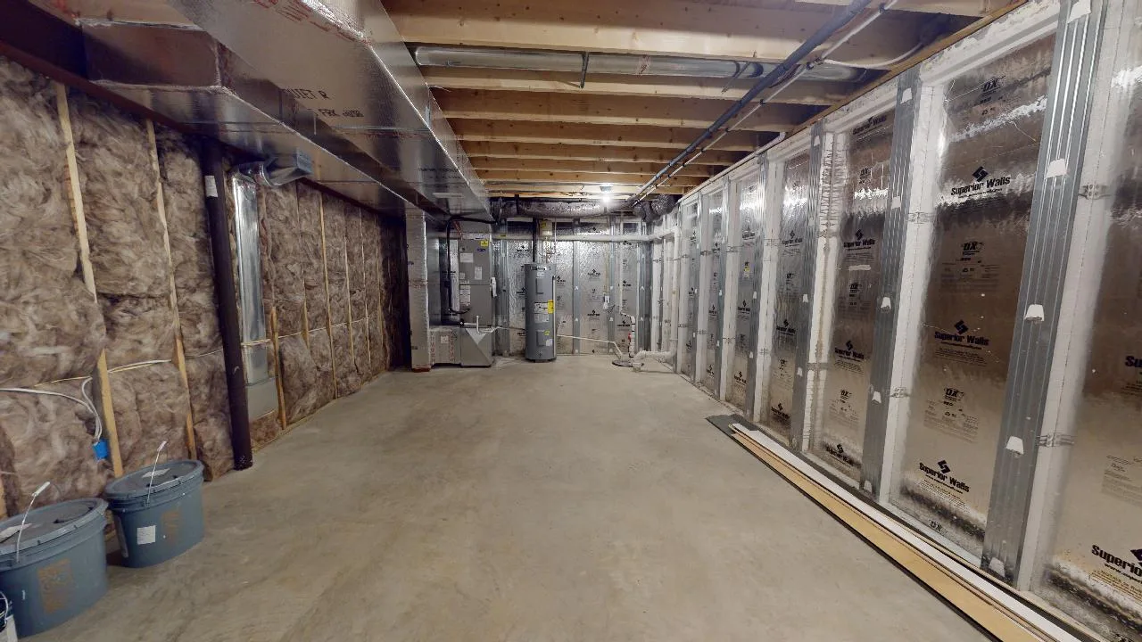 Unfinished basement with exposed wooden ceiling beams, insulated walls, HVAC system, and concrete floor. Construction materials and buckets are placed along the walls. Bright lighting illuminates the space.