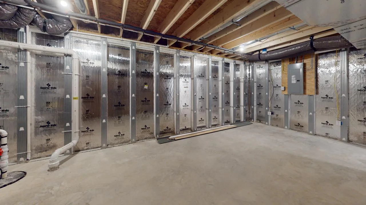 Unfinished basement with exposed ceiling beams, insulation panels on the walls, visible pipes, electrical panel, and a concrete floor. Some wooden planks are stacked against the wall.