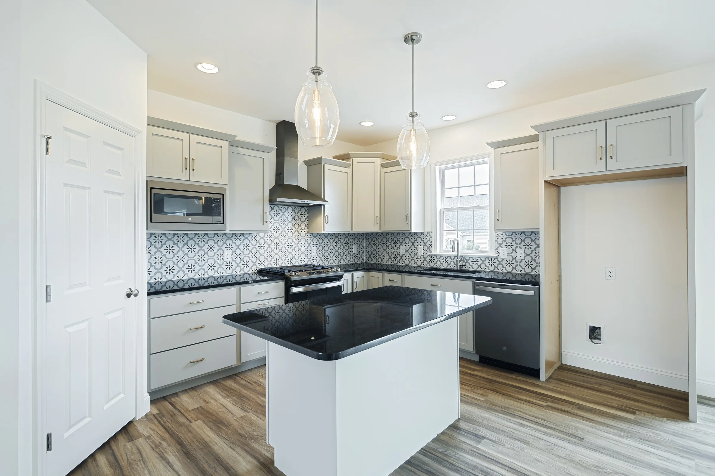 Modern kitchen with light gray cabinets, black countertops, patterned tile backsplash, stainless steel appliances, a center island, wood flooring, pendant lights, and an empty nook for a refrigerator.