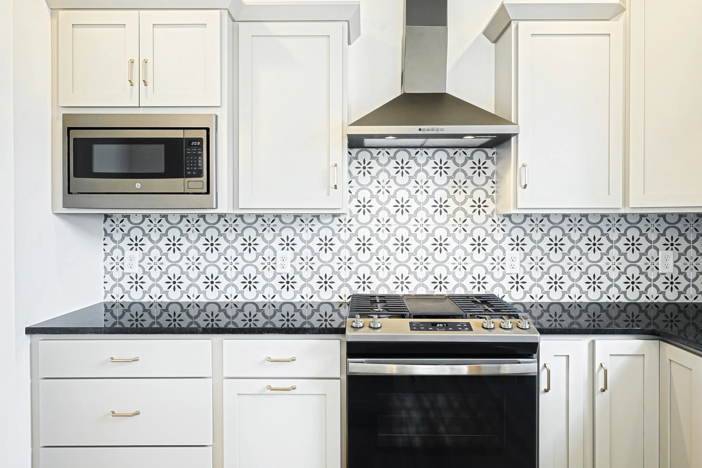 Modern kitchen with white cabinets, a black countertop, stainless steel oven and hood, built-in microwave, and a black-and-white patterned tile backsplash.