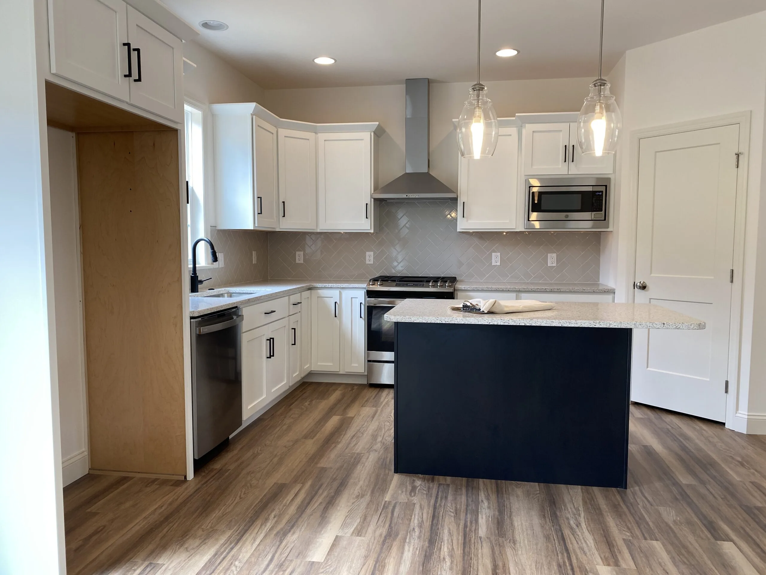 Modern kitchen with white cabinets, stainless steel appliances, a black island with granite countertop, pendant lights, light gray tile backsplash, and wood-look flooring. An empty alcove for a refrigerator is on the left.