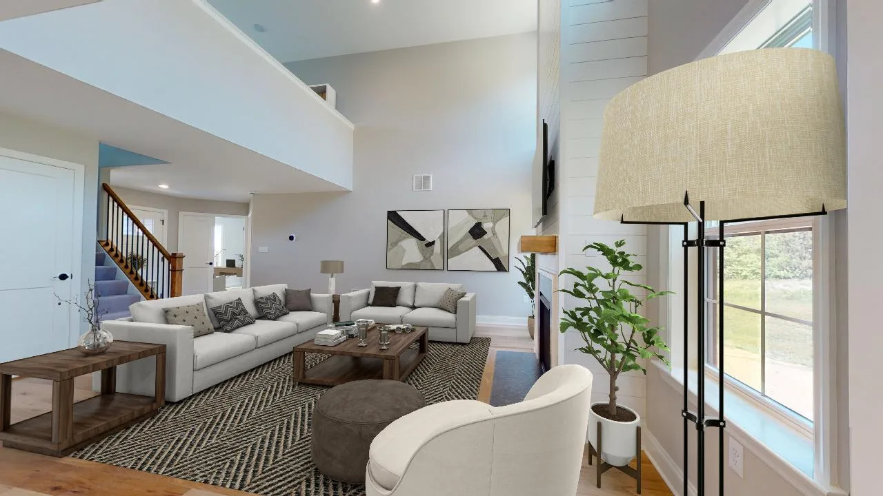 A modern living room with neutral-toned sofas, a patterned rug, a round ottoman, floor lamp, indoor plant, and large windows letting in natural light. Abstract art hangs on the wall above the couch.