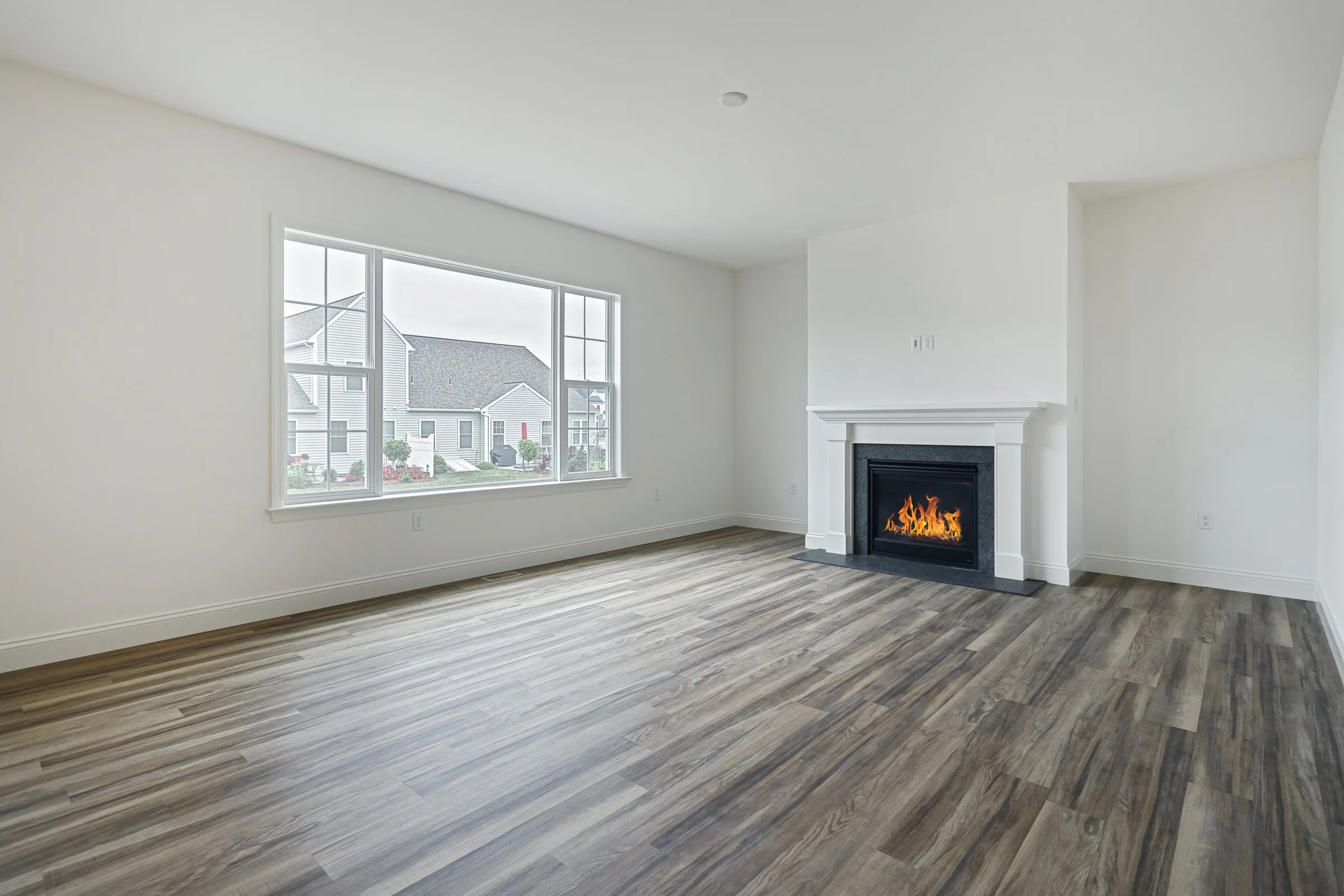 A bright, empty living room with wood flooring, a large window overlooking houses, and a white fireplace with a lit fire. The walls and ceiling are painted white, creating a clean and modern look.
