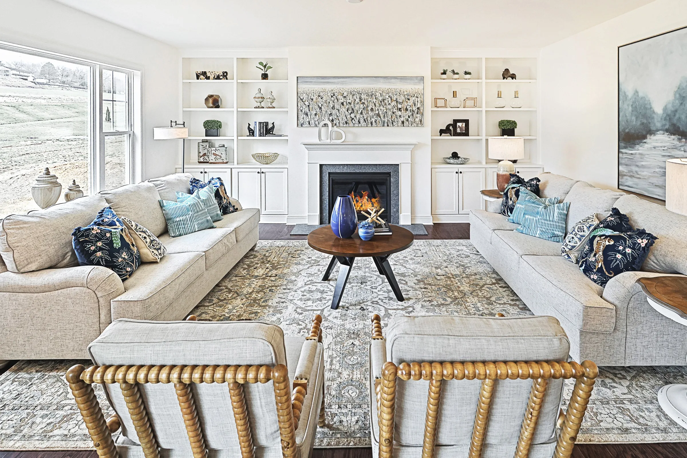 A bright, elegant living room with two beige sofas, two wooden chairs, a central round coffee table, a patterned rug, and a fireplace with built-in shelves and decor. Large windows let in natural light.