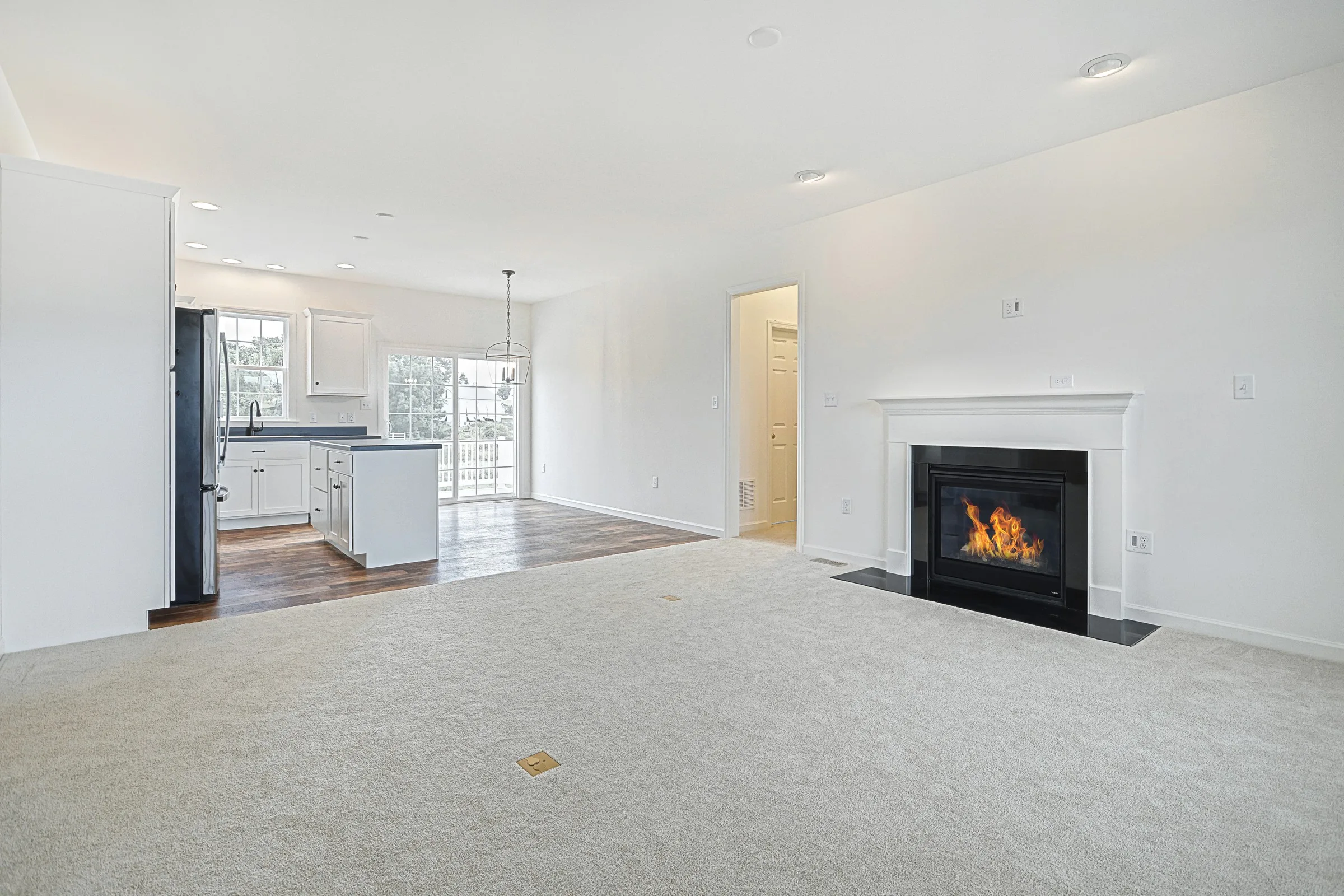 A bright, open-concept living area with light carpet, a black-framed fireplace, and an adjoining kitchen featuring white cabinets, dark flooring, and large windows letting in natural light.
