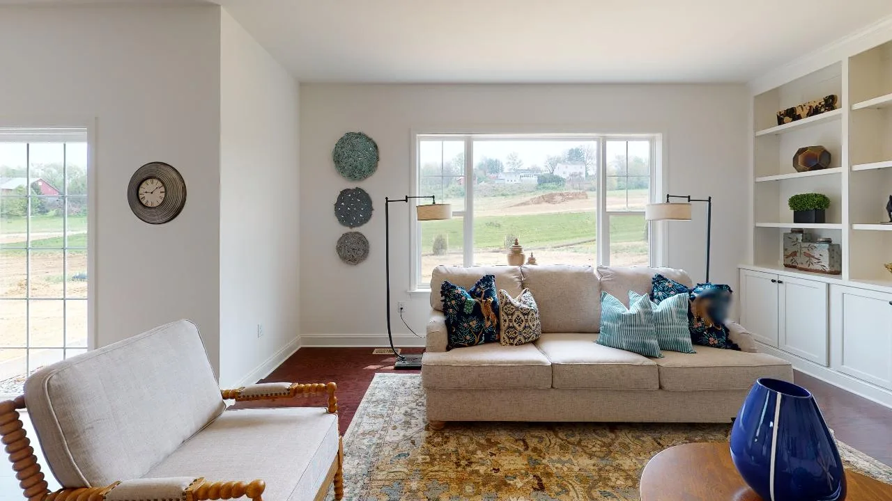 A cozy living room with a beige sofa and armchair, colorful pillows, a blue vase on a wooden table, wall art, built-in shelves, and large windows overlooking a grassy landscape.