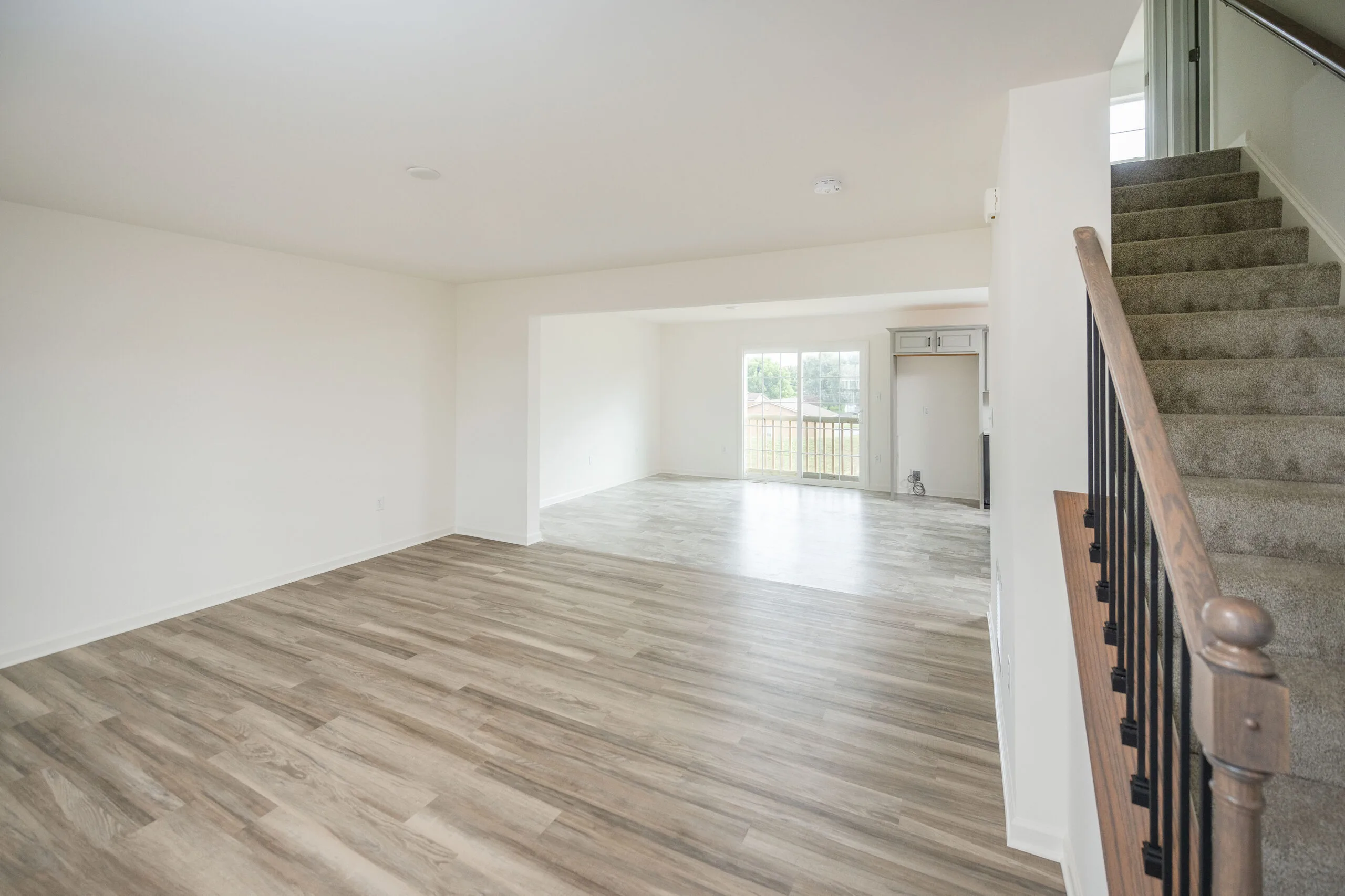Bright, empty room with light wood flooring, white walls, and a staircase with beige carpet and a wooden handrail. Large windows in the background let in natural light, illuminating the open living space.