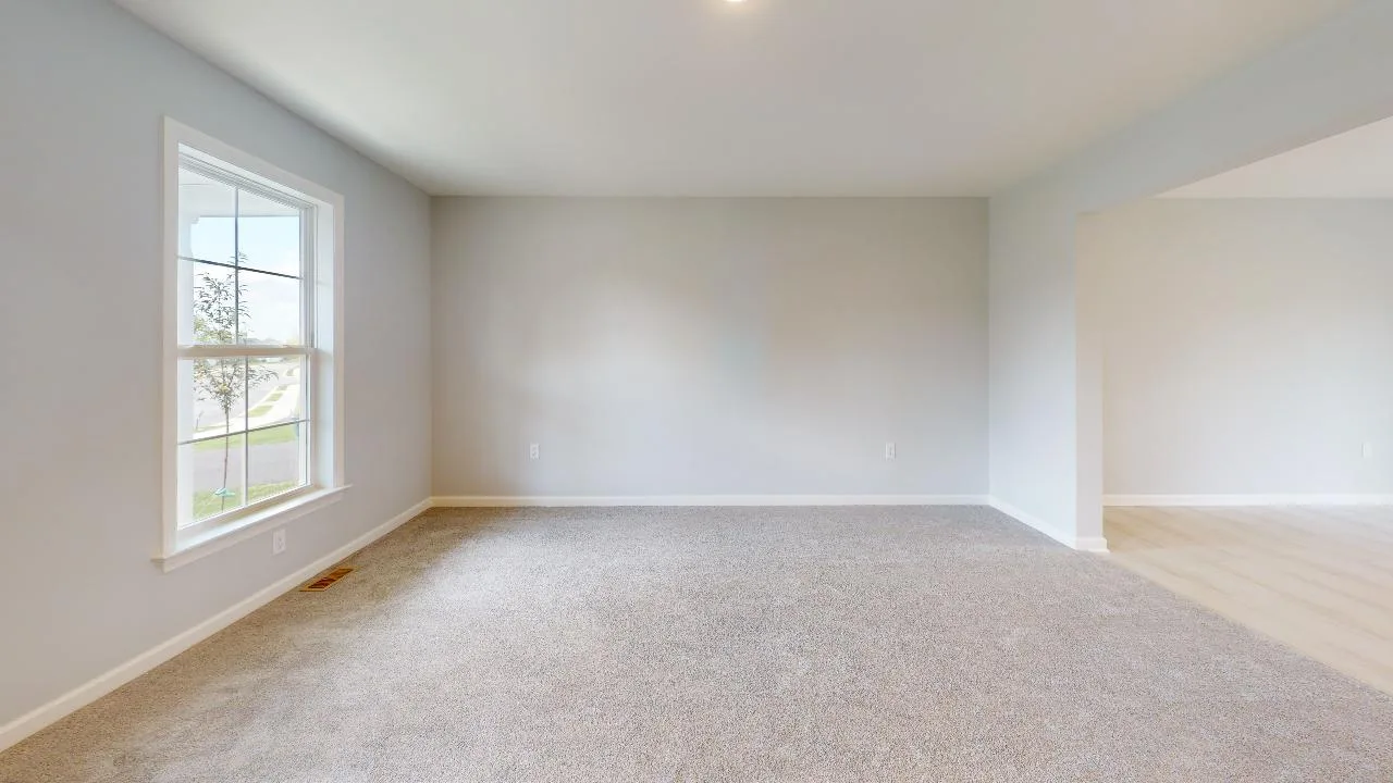 A bright, empty room with light gray carpet, pale gray walls, a large window on the left letting in natural light, and an open doorway leading to a space with light-colored flooring.