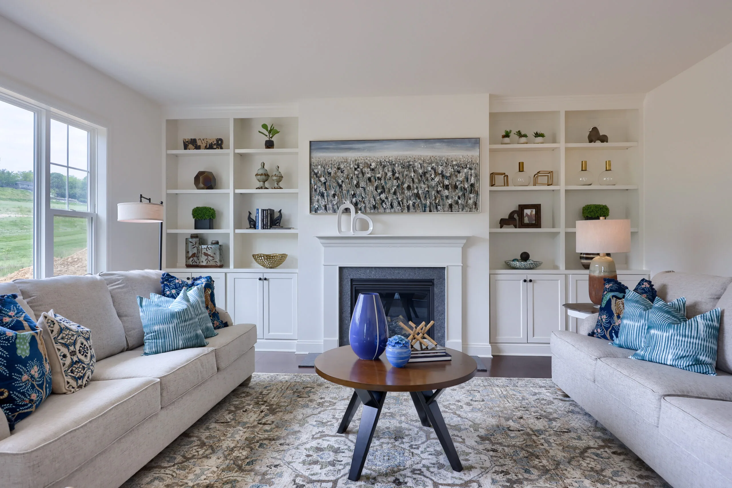 Bright living room with two beige sofas, blue accent pillows, a round wooden coffee table, a patterned rug, built-in white shelves, decor items, a fireplace, and a large wall art above the mantel. Large windows let in natural light.