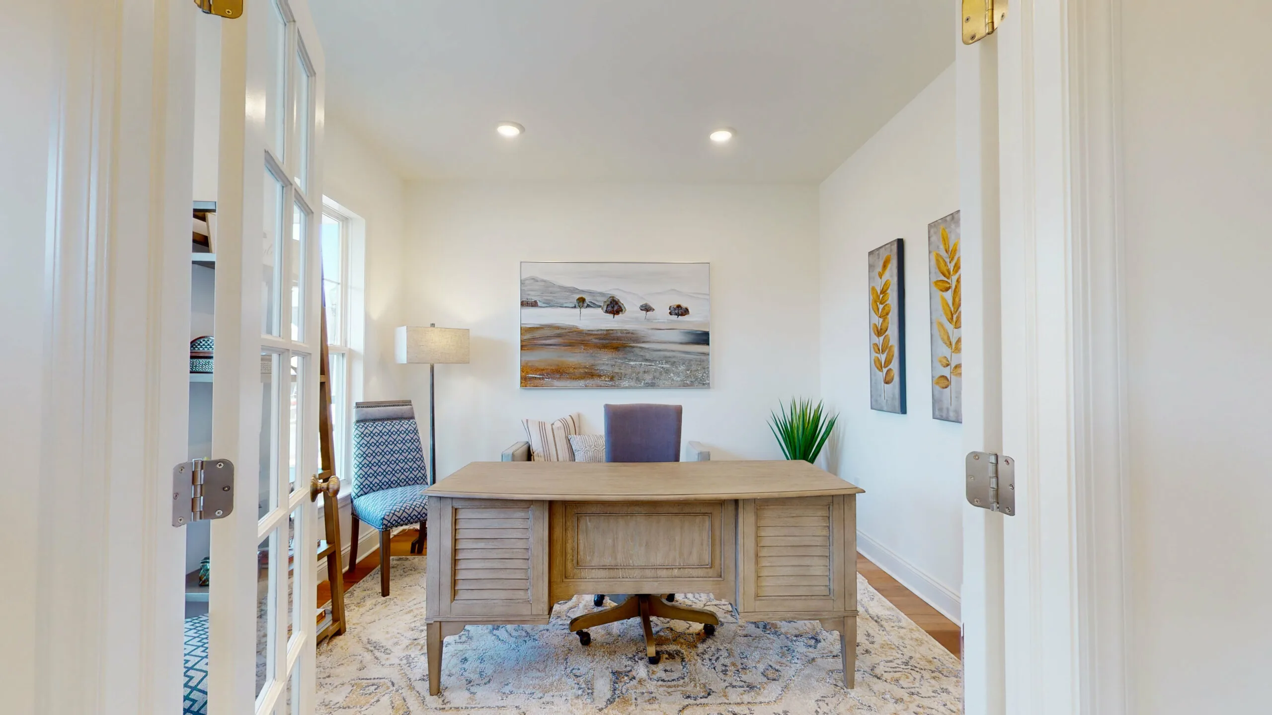 A bright home office with a wooden desk, gray chair, two accent chairs, a floor lamp, wall art, and a potted plant. The room has white walls, a patterned rug, and double glass doors.