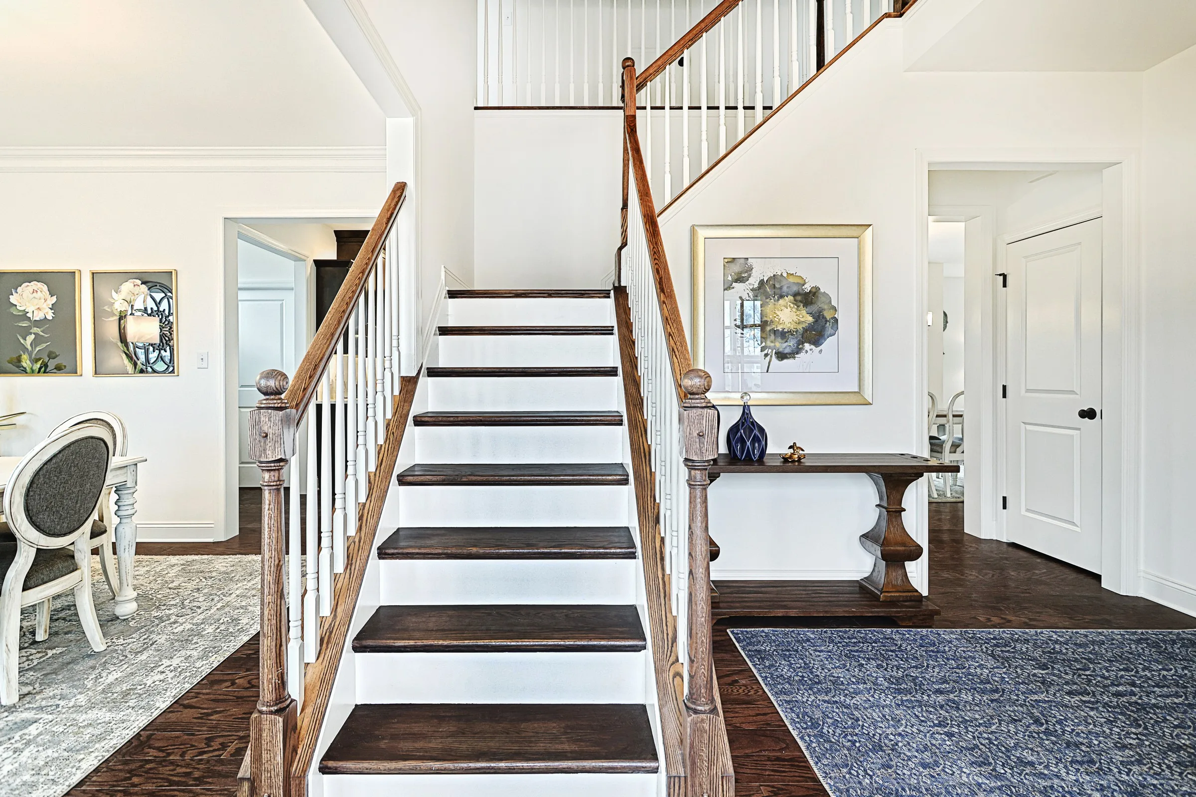 A bright, modern entryway with a wooden staircase featuring white risers and dark treads, a blue rug, framed artwork, and a view into a dining area with elegant chairs and floral wall art.