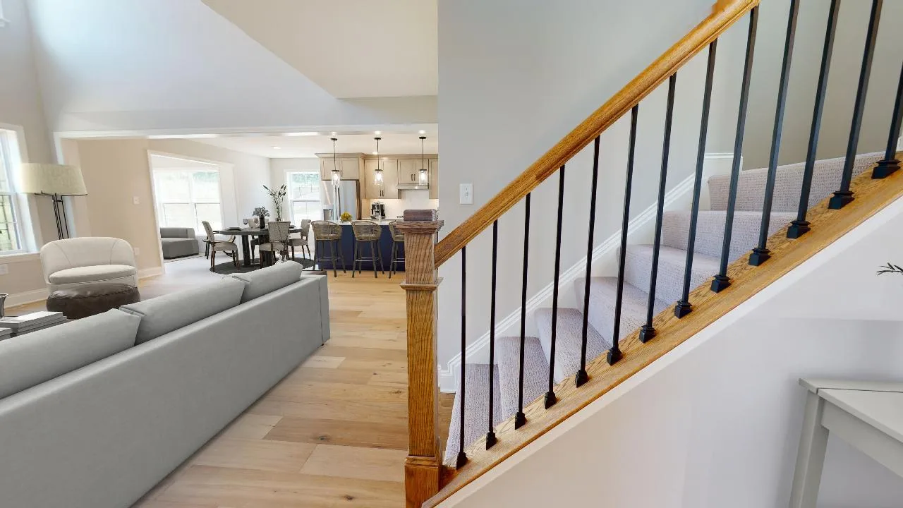 A modern open-concept living area with a light gray sofa, dining table, and kitchen in the background. Carpeted stairs with a wooden handrail and black spindles are visible in the foreground. Natural light fills the space.