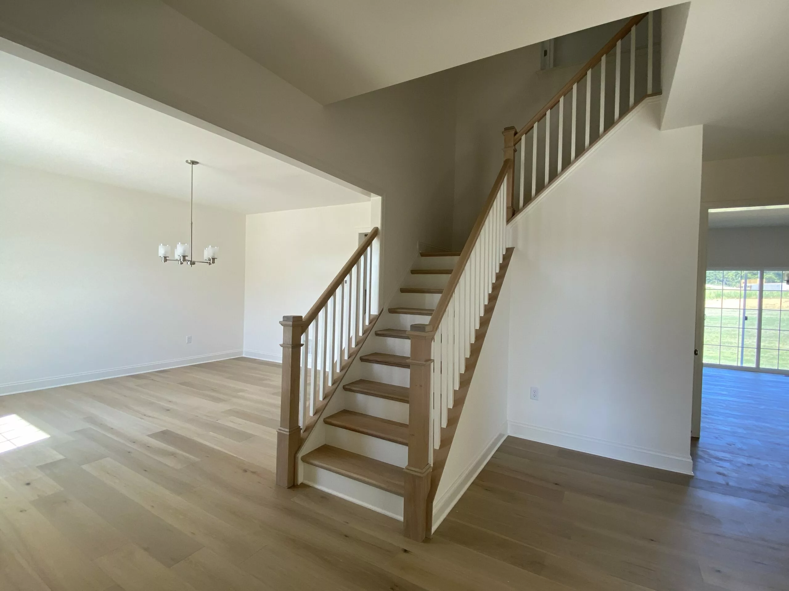 A bright, empty room with light wood floors, white walls, a wooden staircase with white railings, a six-light chandelier, and sliding glass doors leading outside.