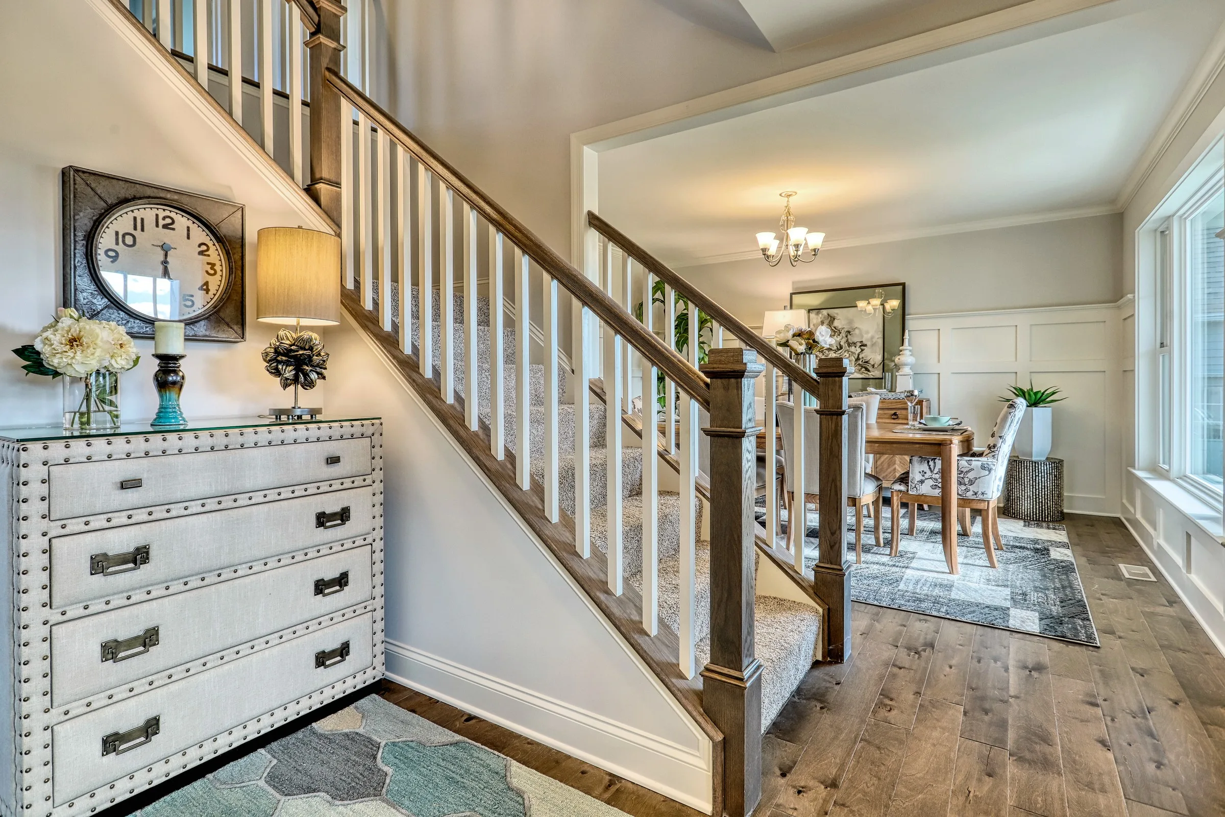 A modern entryway with a wooden staircase, a white chest with nailhead trim, a round clock, flowers, and a lamp. Adjacent dining area features a wooden table, chairs, a chandelier, and large windows.