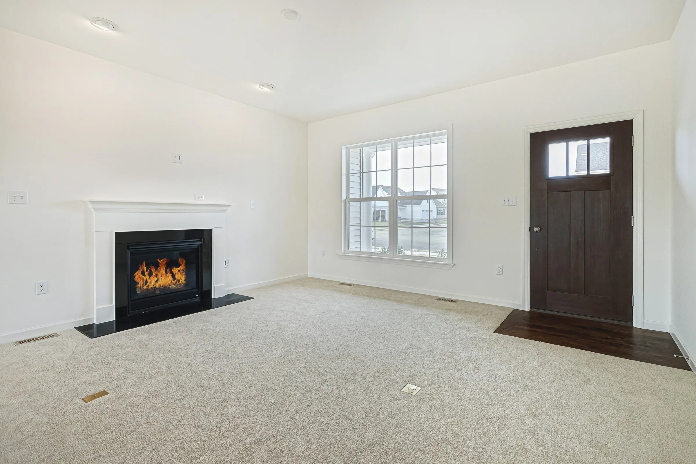 A bright, empty living room with cream-colored carpet, white walls, a lit fireplace, a large window, and a dark wooden front door.