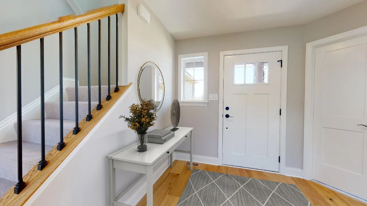 Bright entryway with light wood floors, white front door, a white console table with a round mirror, books, vase, and plant. Carpeted stairs with black railings are on the left; gray rug on the floor.