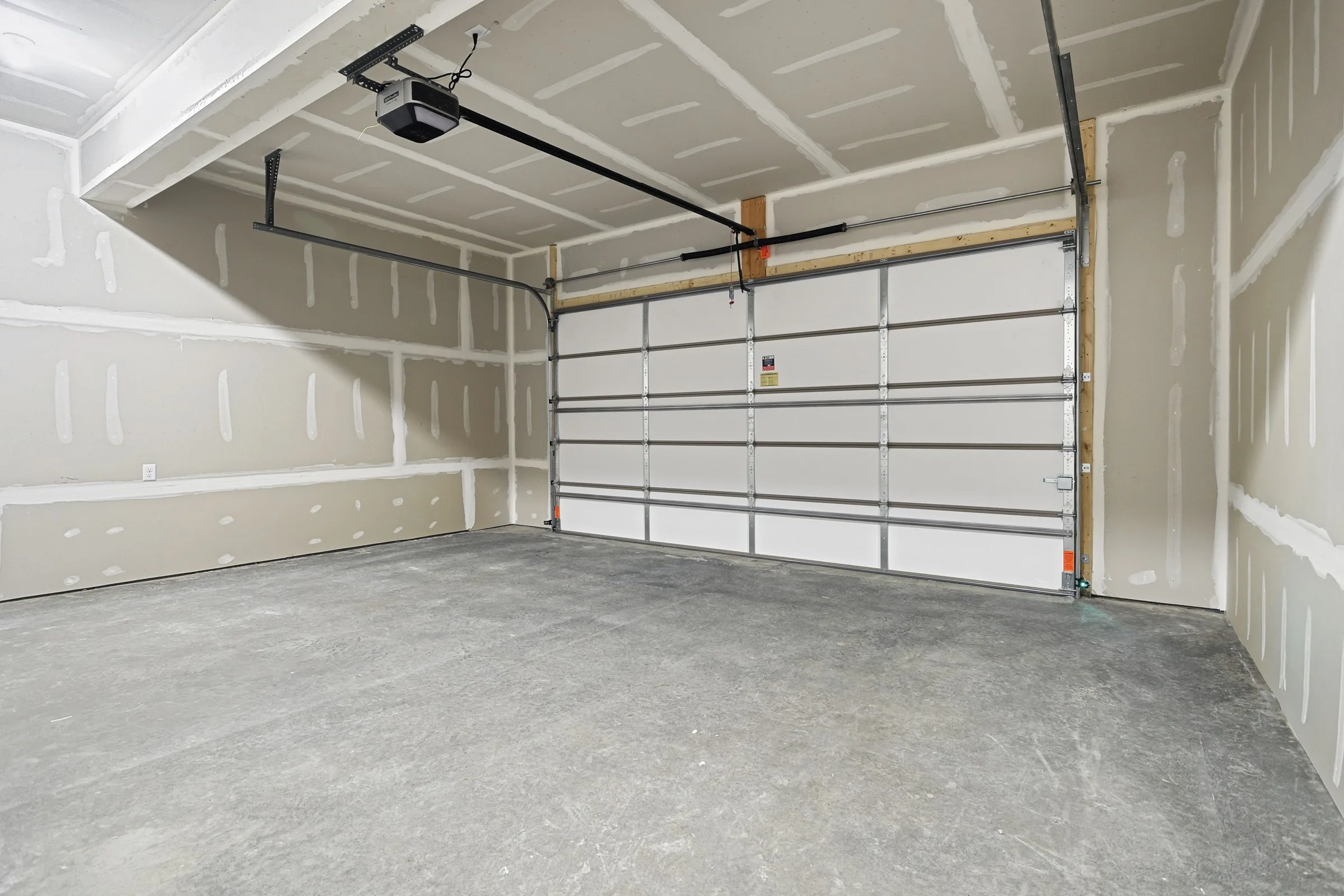 An empty, unfinished two-car garage with exposed drywall, a concrete floor, and a closed automatic overhead garage door. The space is clean and well-lit.
