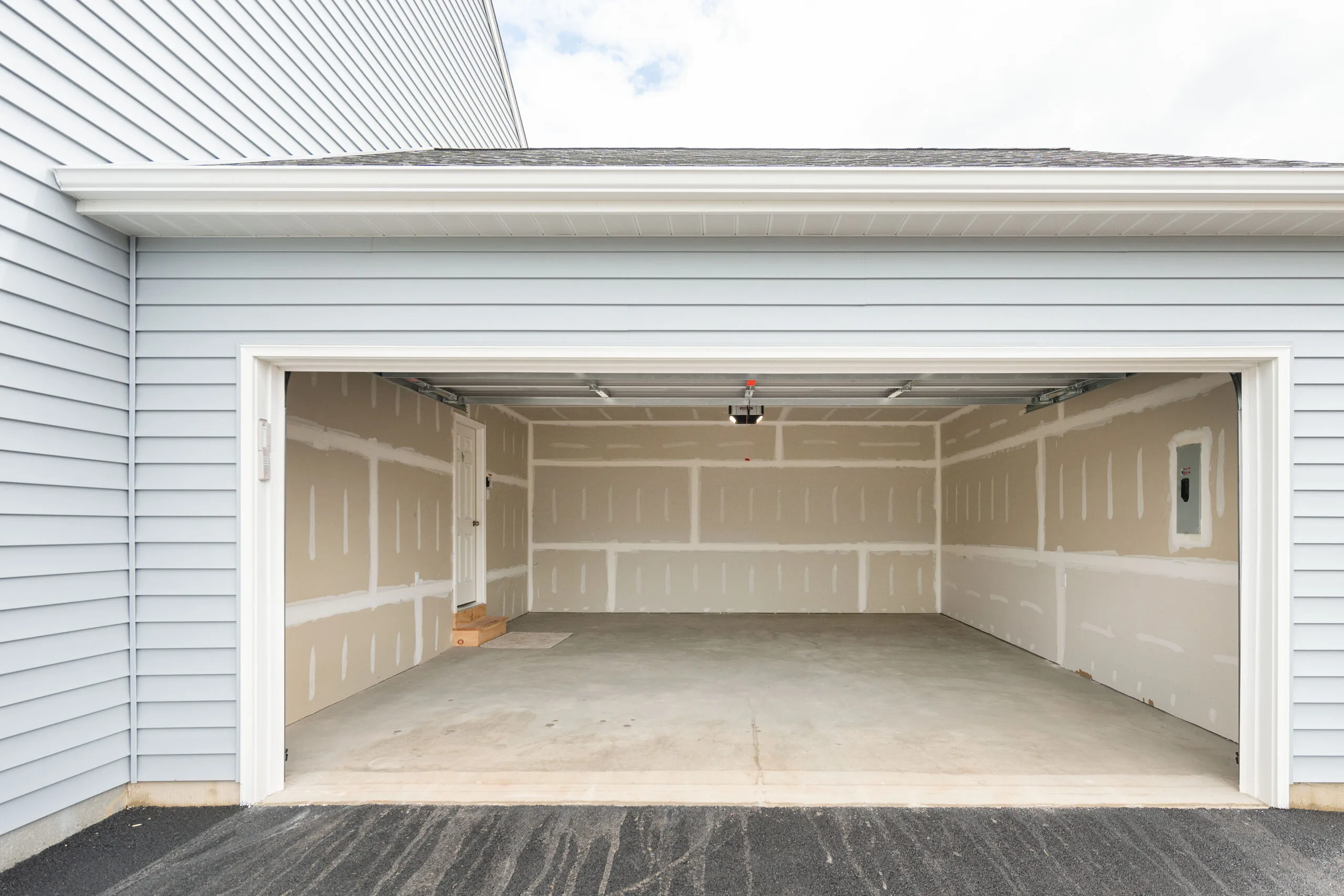 An empty, two-car garage with unfinished drywall walls and a concrete floor, attached to a light blue house with white trim. The garage door is open, revealing the spacious interior.