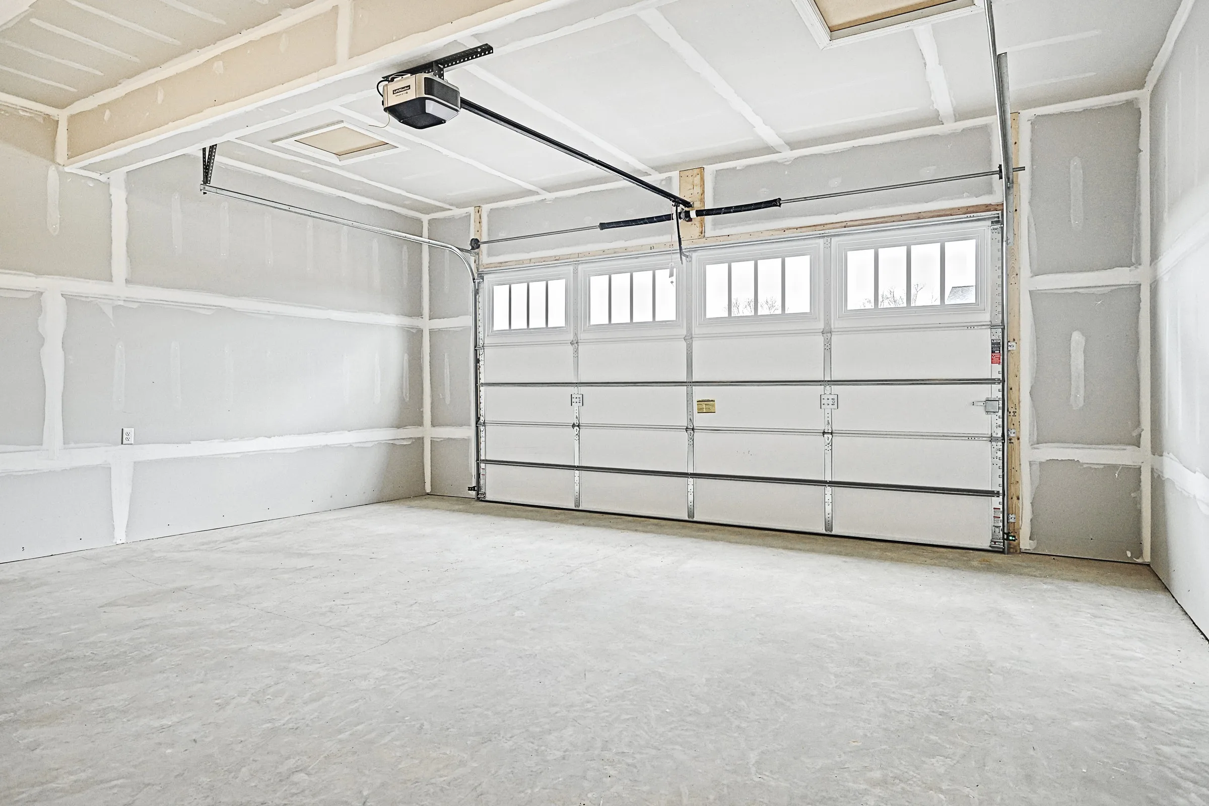 Empty, newly constructed garage with unfinished drywall, concrete floor, and a closed, white double garage door with windows at the top. Ceiling has exposed seams, and a garage door opener is mounted overhead.