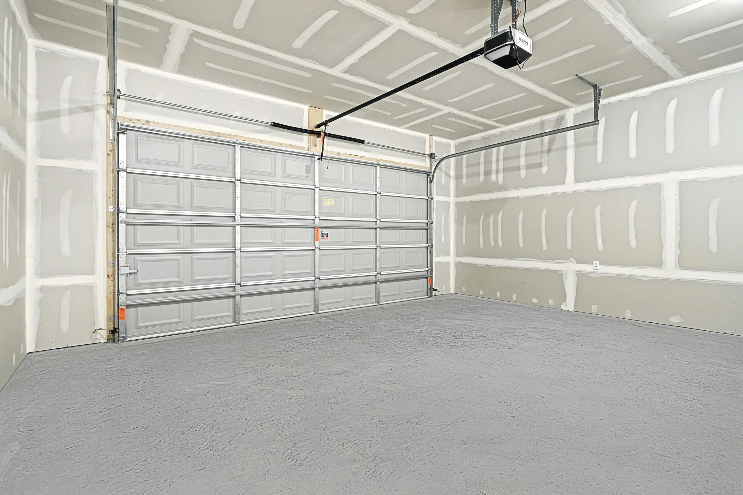 A clean, empty garage with unfinished drywall, a closed double garage door, ceiling-mounted garage door opener, and a smooth gray concrete floor.