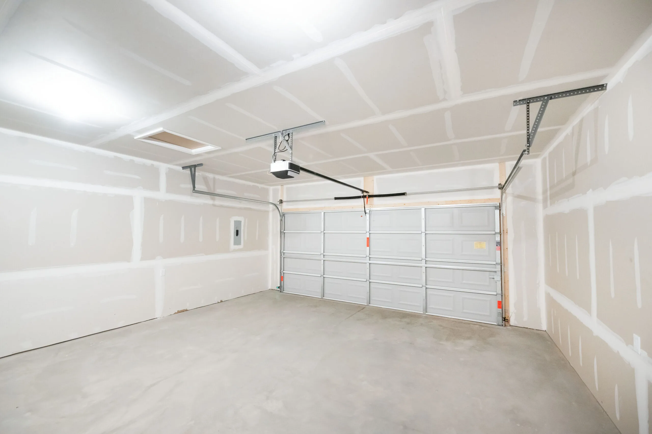 Empty, newly constructed two-car garage with unfinished drywall, concrete floor, ceiling lights, automatic garage door opener, and a double garage door. The space is clean and well-lit.