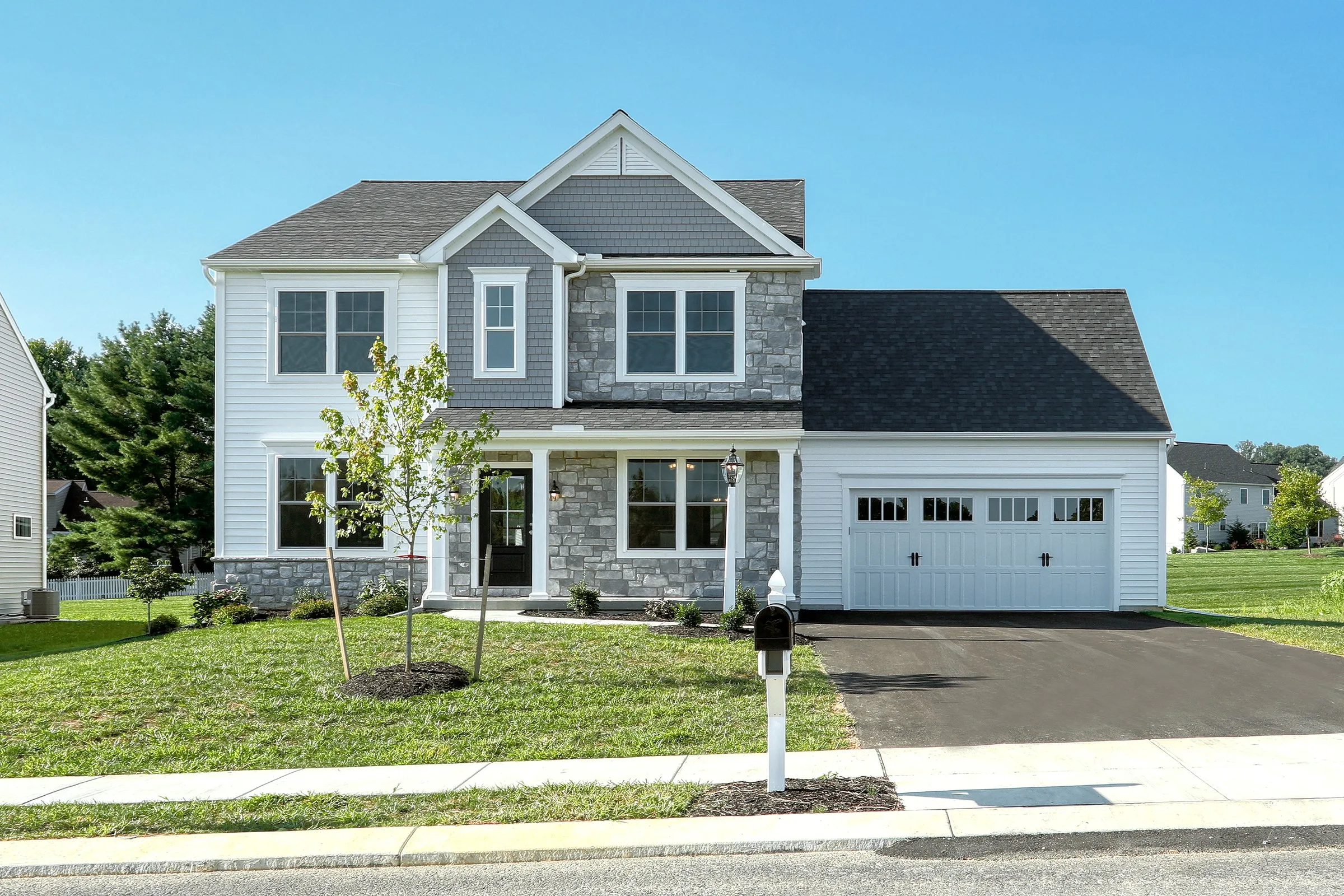 Two-story suburban house with gray stone and white siding exterior, black roof, double garage, front lawn, small trees, and a white mailbox by the driveway, on a clear sunny day.