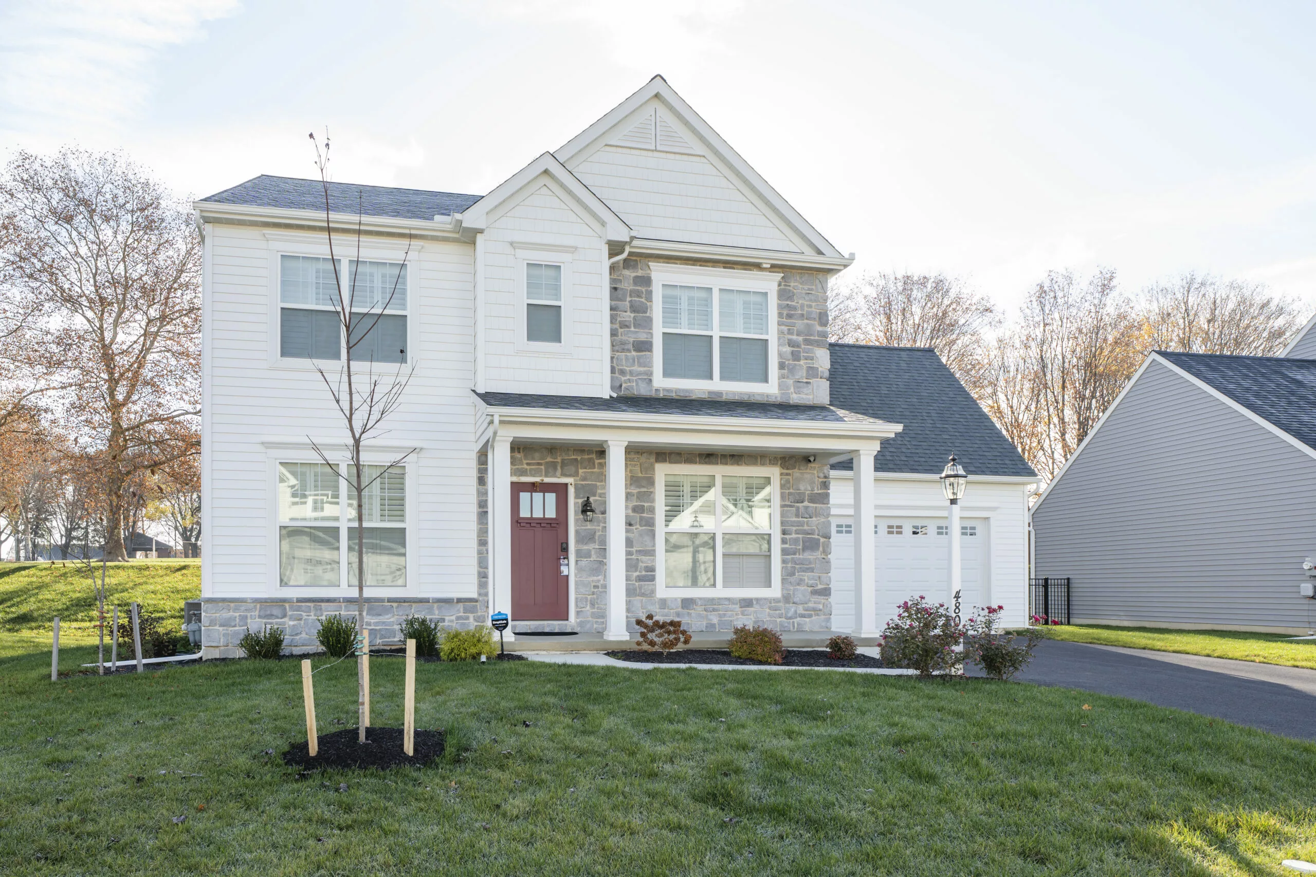 A two-story suburban house with white siding, stone accents, a red front door, and attached double garage, surrounded by a neatly landscaped yard and a small tree in the front.