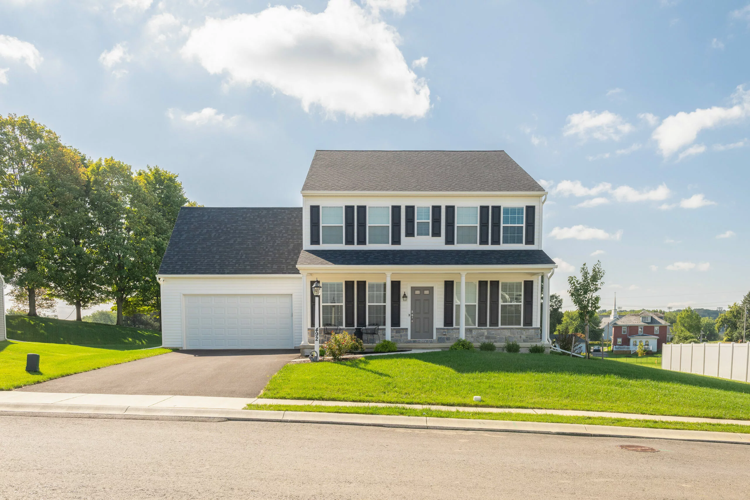 A two-story suburban house with white siding, black shutters, and a gray roof. It has a front porch, attached garage, and is surrounded by a well-kept green lawn under a sunny sky with some clouds.