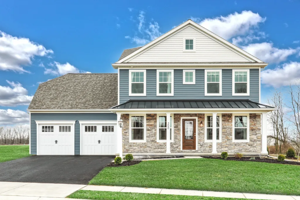 A modern two-story house with blue siding, stone accents, and a covered front porch. It has white-trimmed windows, double garage doors, a black driveway, and a well-manicured lawn under a partly cloudy sky.