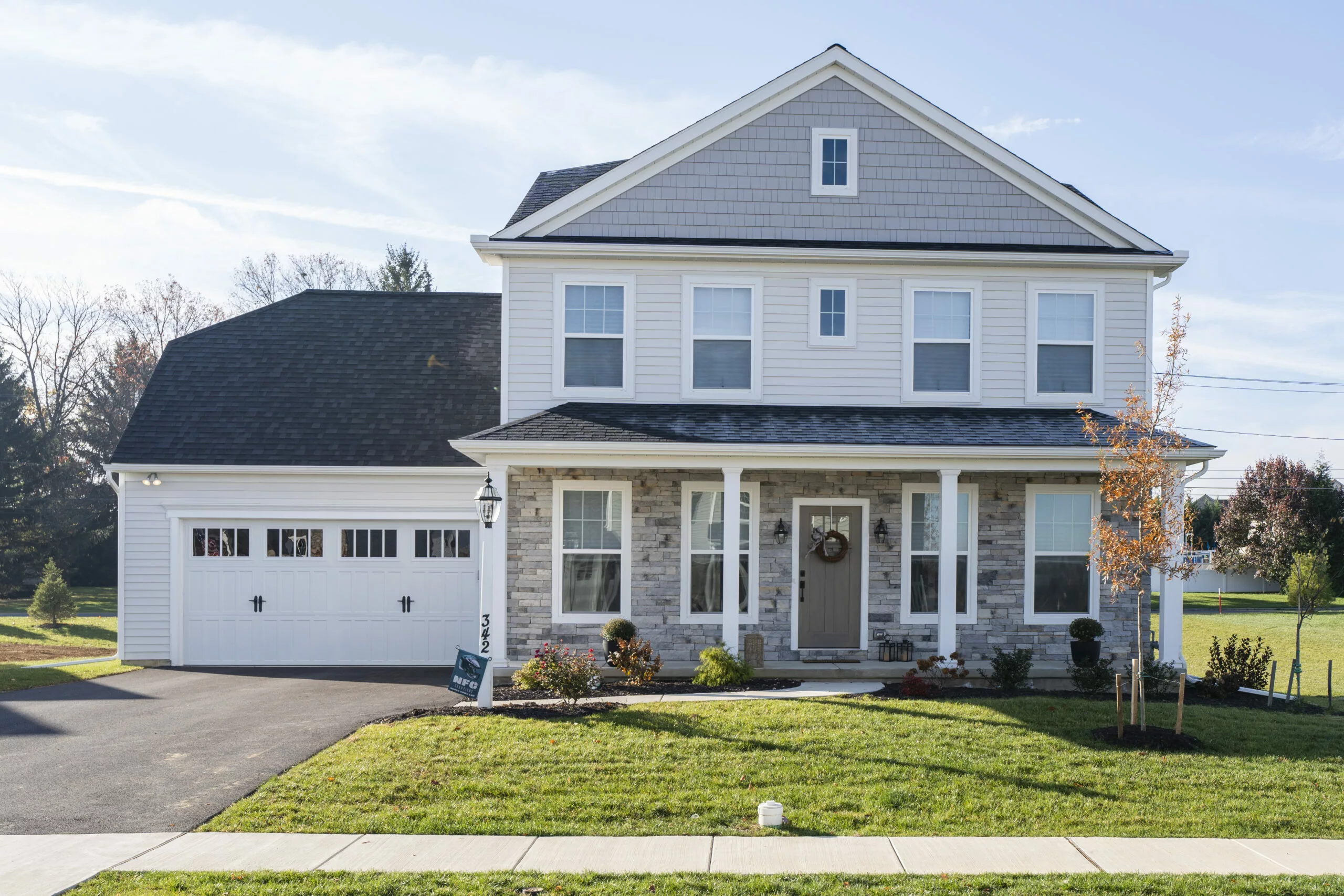 A two-story suburban house with light-colored siding, stone accents, a covered front porch, attached two-car garage, and a well-maintained lawn on a sunny day.