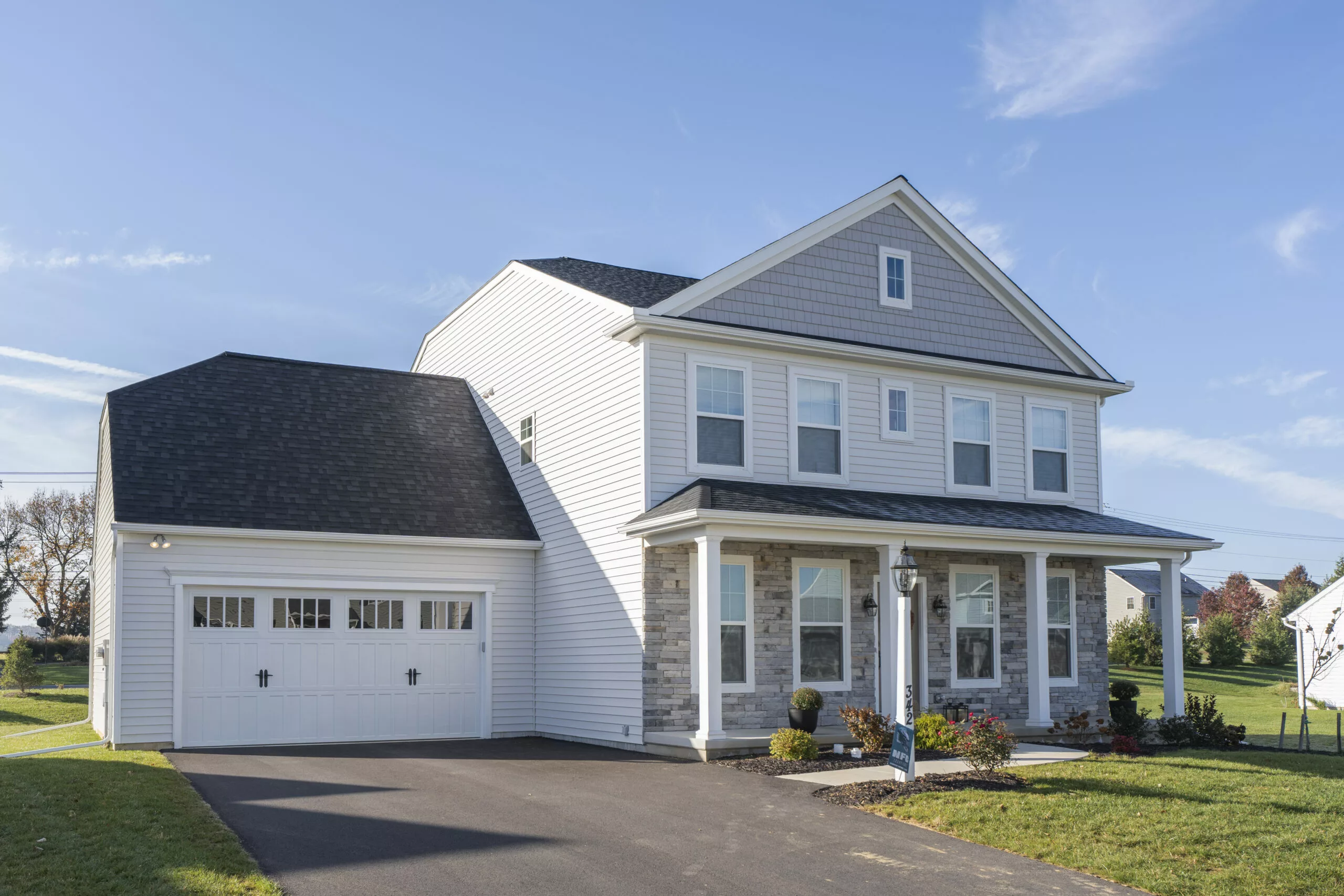 Two-story suburban house with light gray siding, front porch, attached double garage, and neatly landscaped yard on a clear, sunny day.