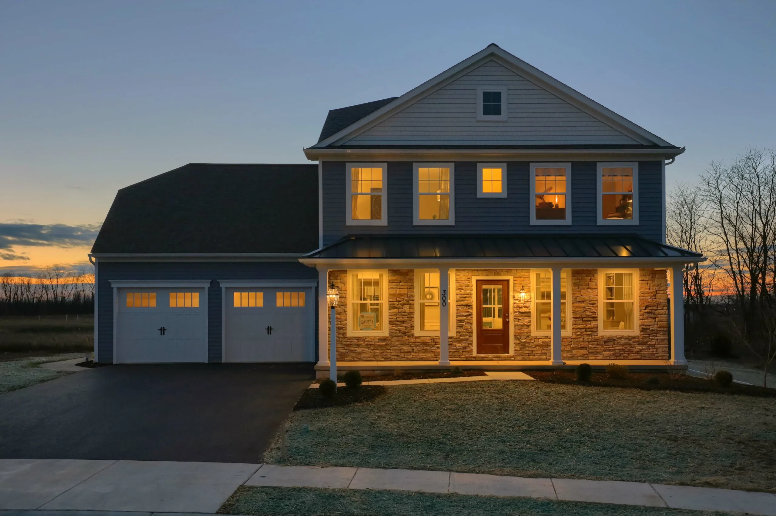 A two-story house with lights on inside at dusk, featuring a front porch, stone accents, and a two-car garage, with a driveway and sidewalk visible in front.