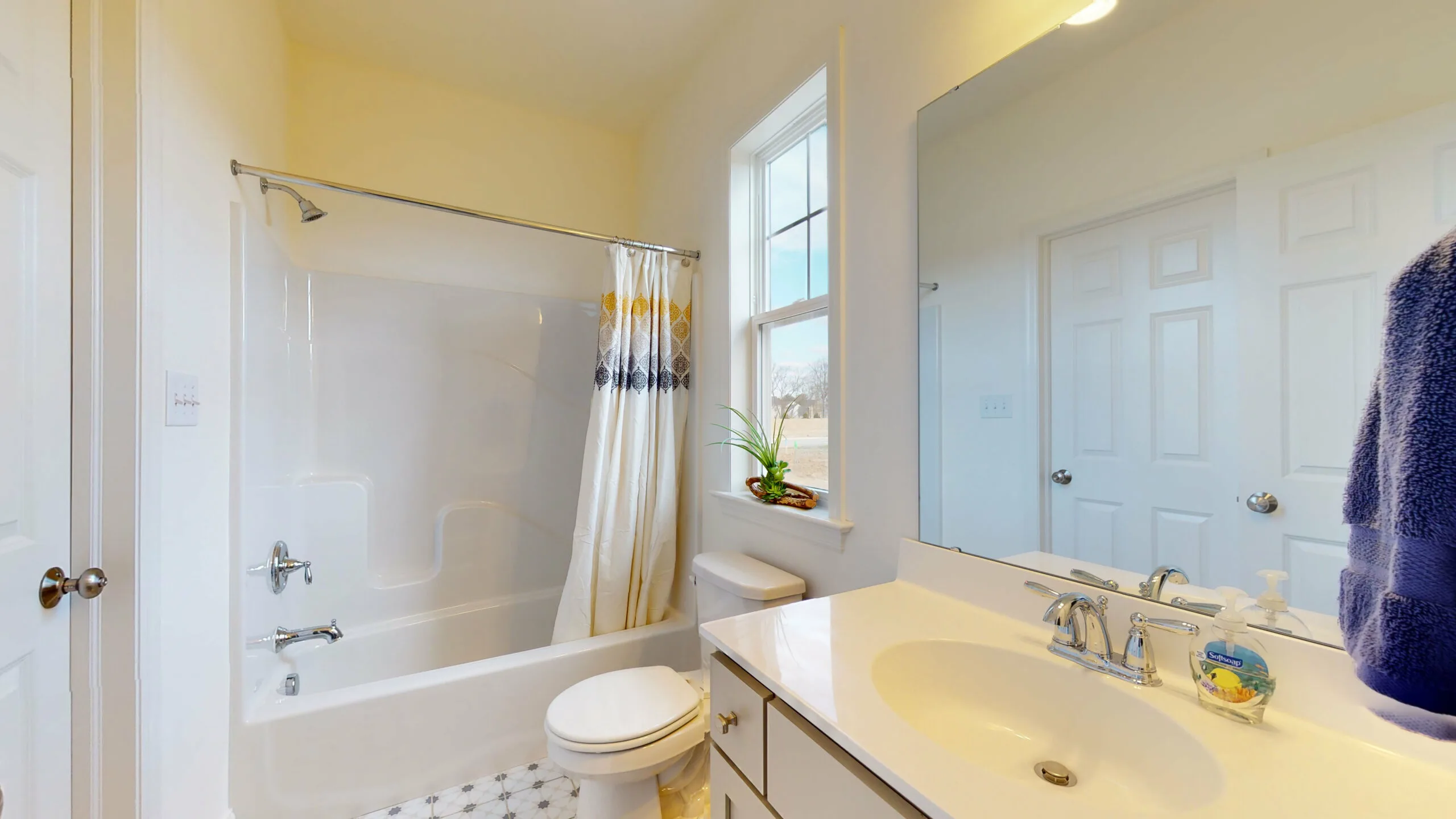 A bright, clean bathroom with a white bathtub and shower, a patterned shower curtain, a toilet, a sink with a large mirror, a window, and a small plant on the windowsill. Blue towels hang on a wall hook.