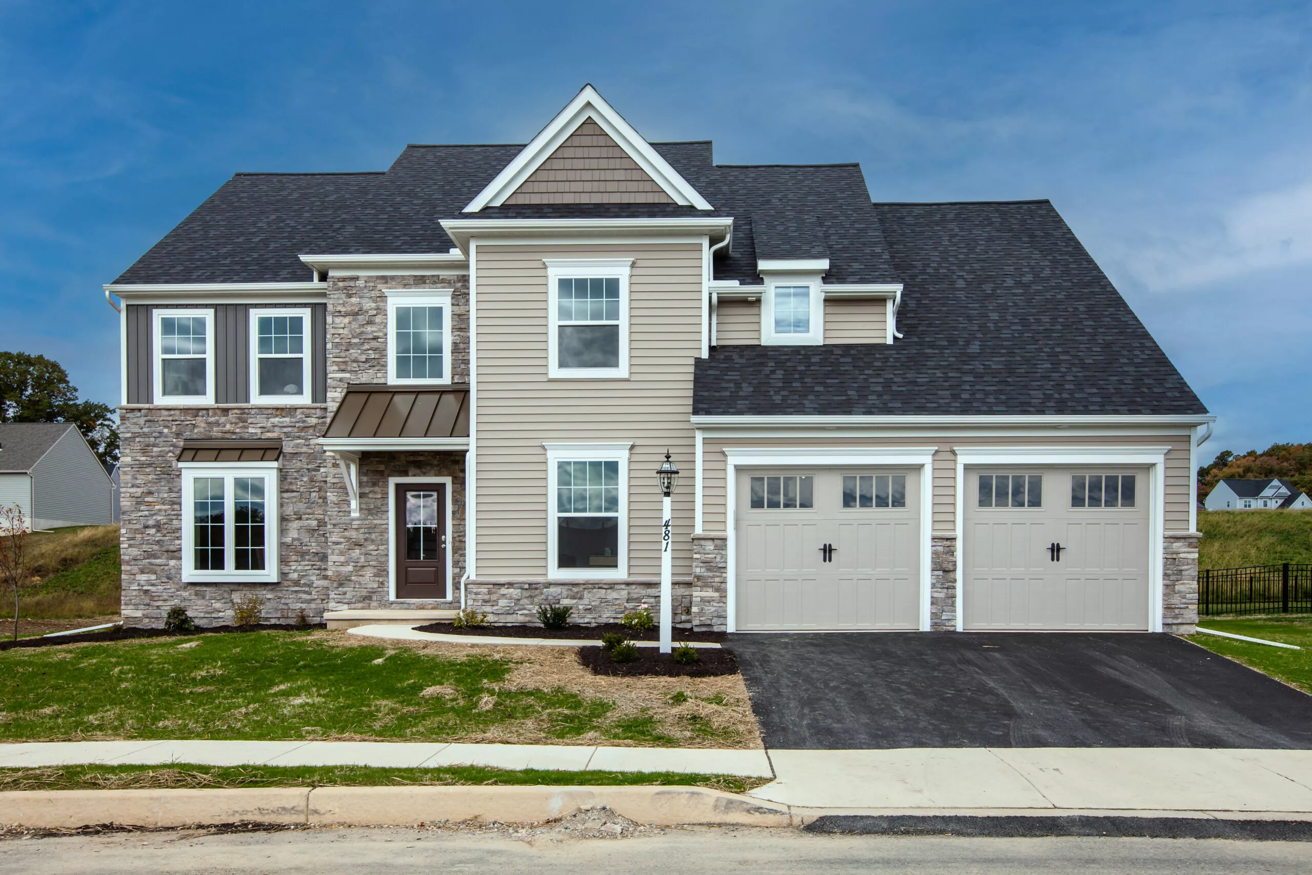 A modern two-story suburban house with stone and beige siding, dark roof shingles, large windows, and a double garage. A driveway and small lawn are in front, with a clear blue sky overhead.