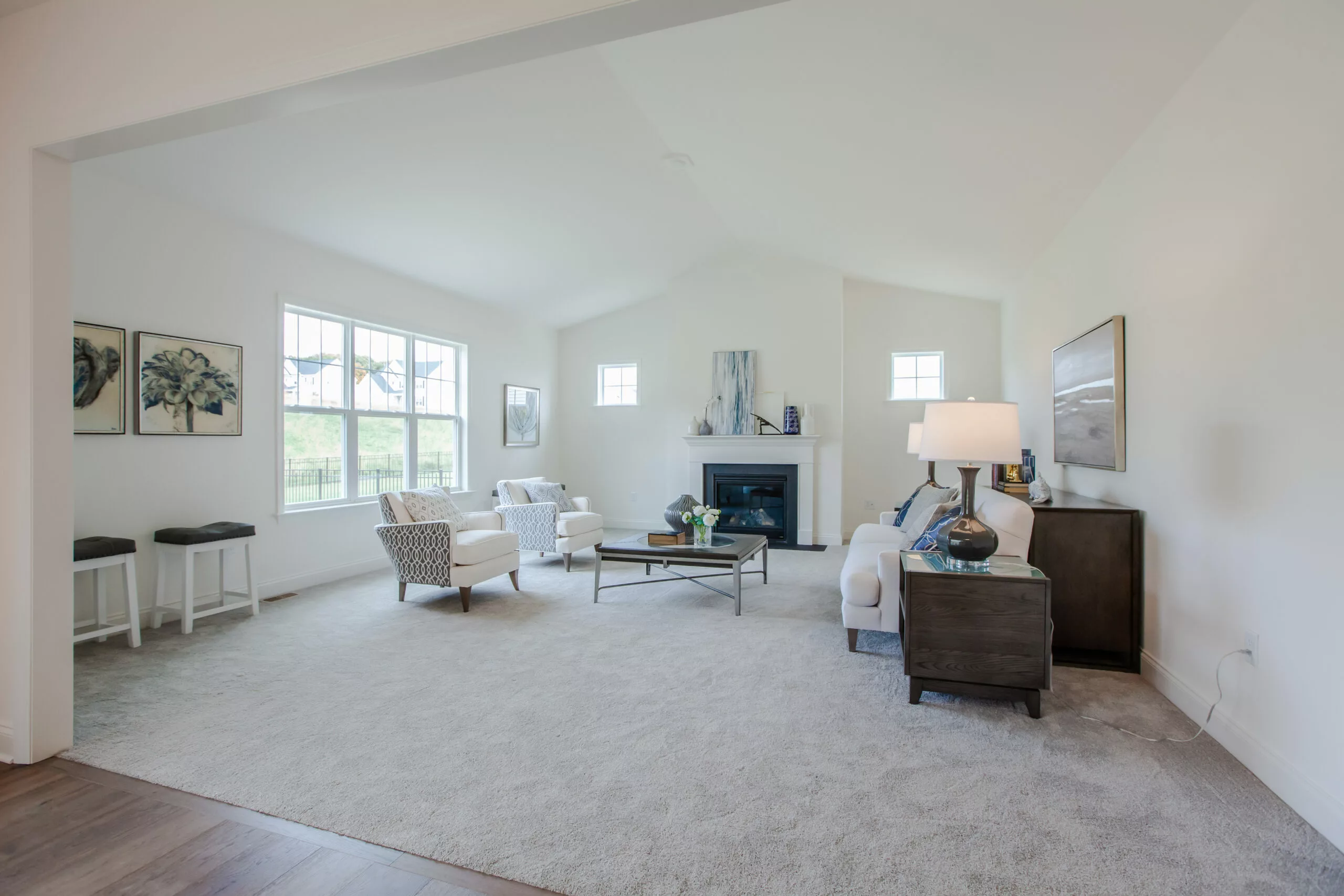 A bright living room with light-colored walls and carpet, featuring large windows, white armchairs, a fireplace, a coffee table, and tasteful decor throughout. Natural light fills the spacious, inviting space.