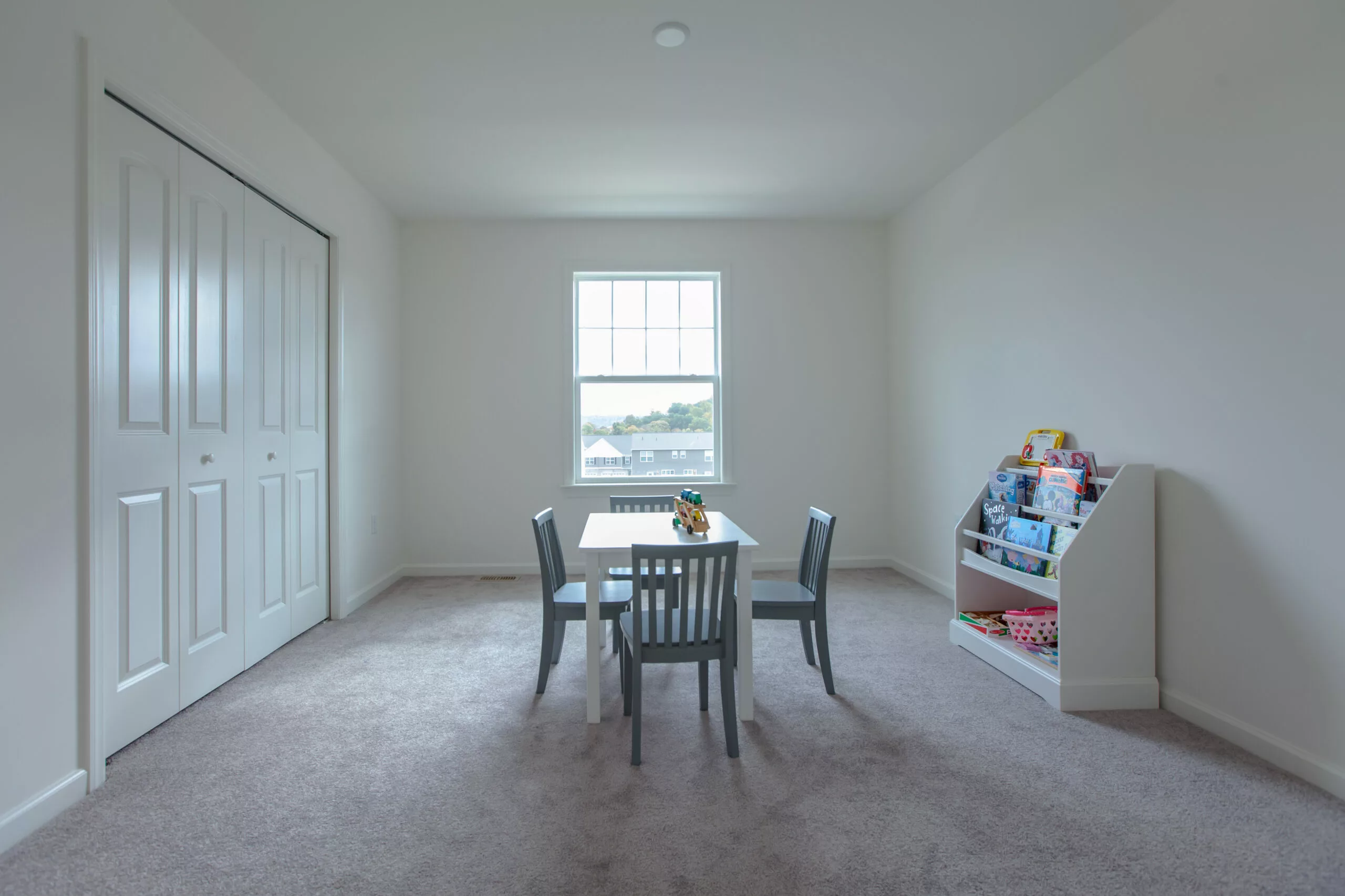 A bright, minimalist room with a small table and three chairs in the center, a window on the far wall, and a bookshelf filled with toys and books to the right on a carpeted floor.