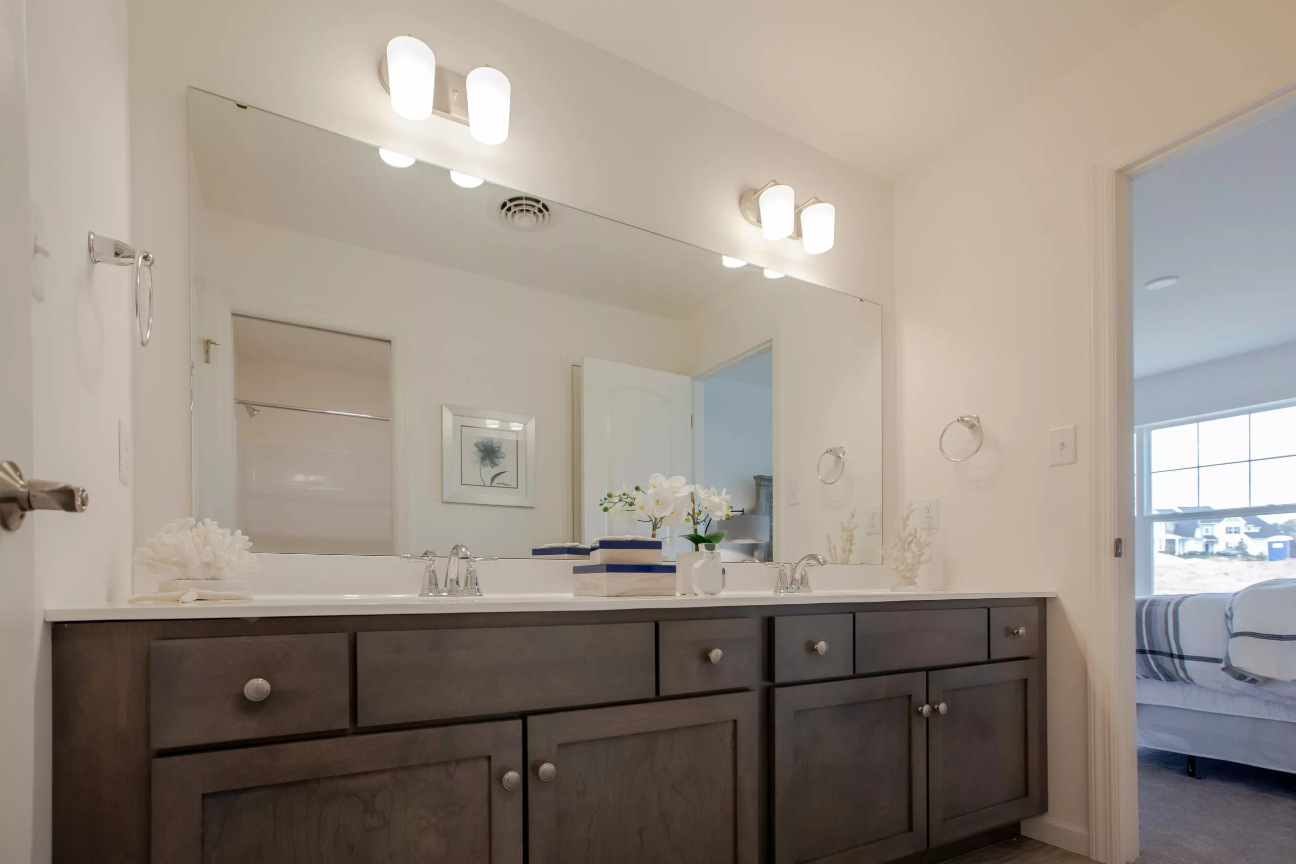 Modern bathroom with a large mirror above a dark wood double-sink vanity, white countertop, two light fixtures, and a view into a bright, minimally furnished bedroom.
