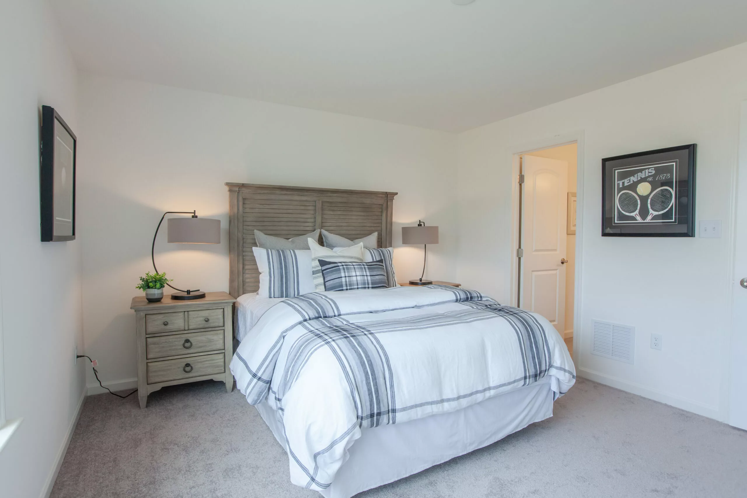 A bright bedroom with a neatly made bed featuring white and gray plaid bedding, a wooden headboard, two bedside tables with lamps, and framed tennis artwork on the wall. A door leads to an adjoining room.