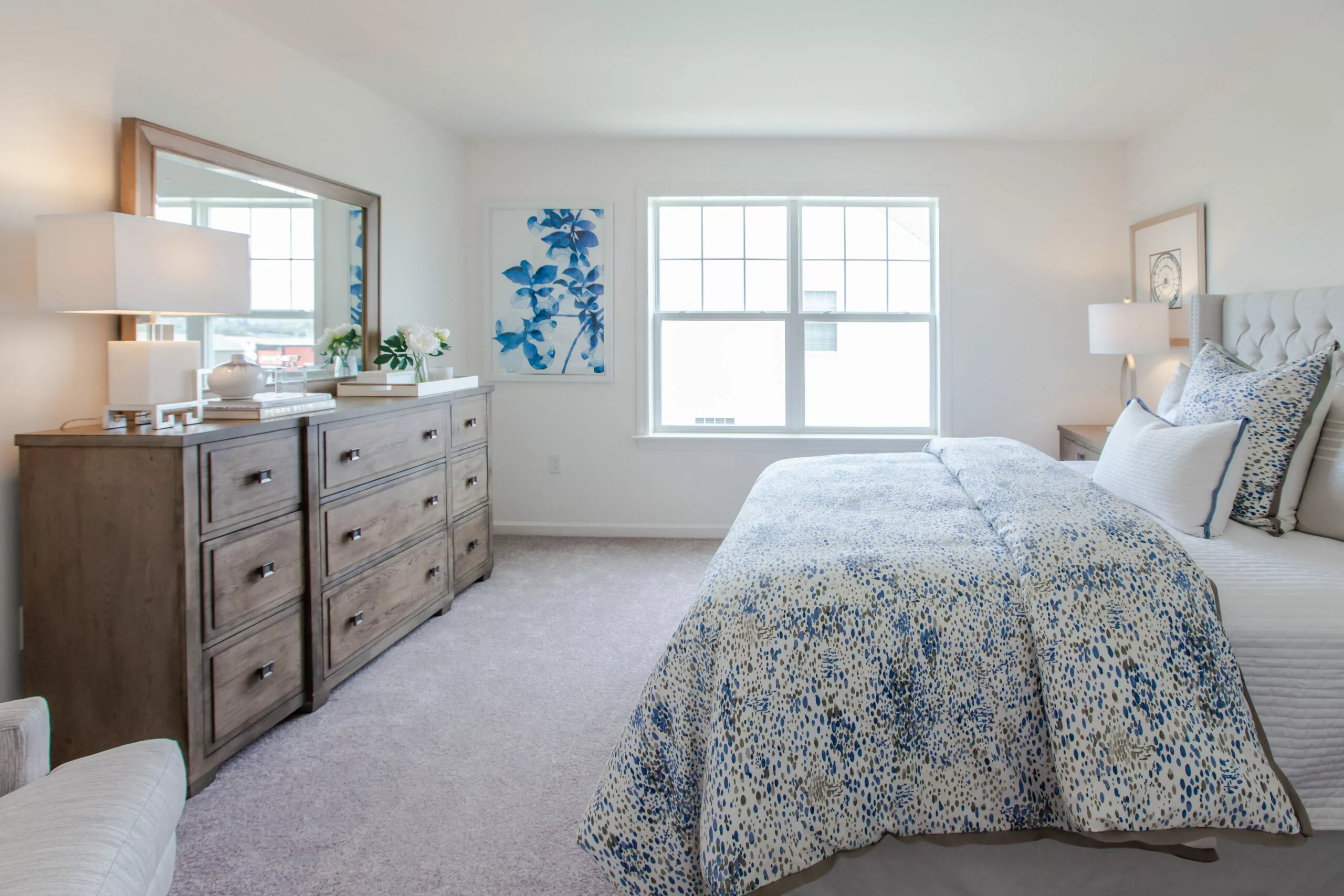 A bright, modern bedroom with a large bed featuring blue and white patterned bedding, a wooden dresser with a mirror, lamps, and plants, a window letting in natural light, and a blue floral artwork on the wall.
