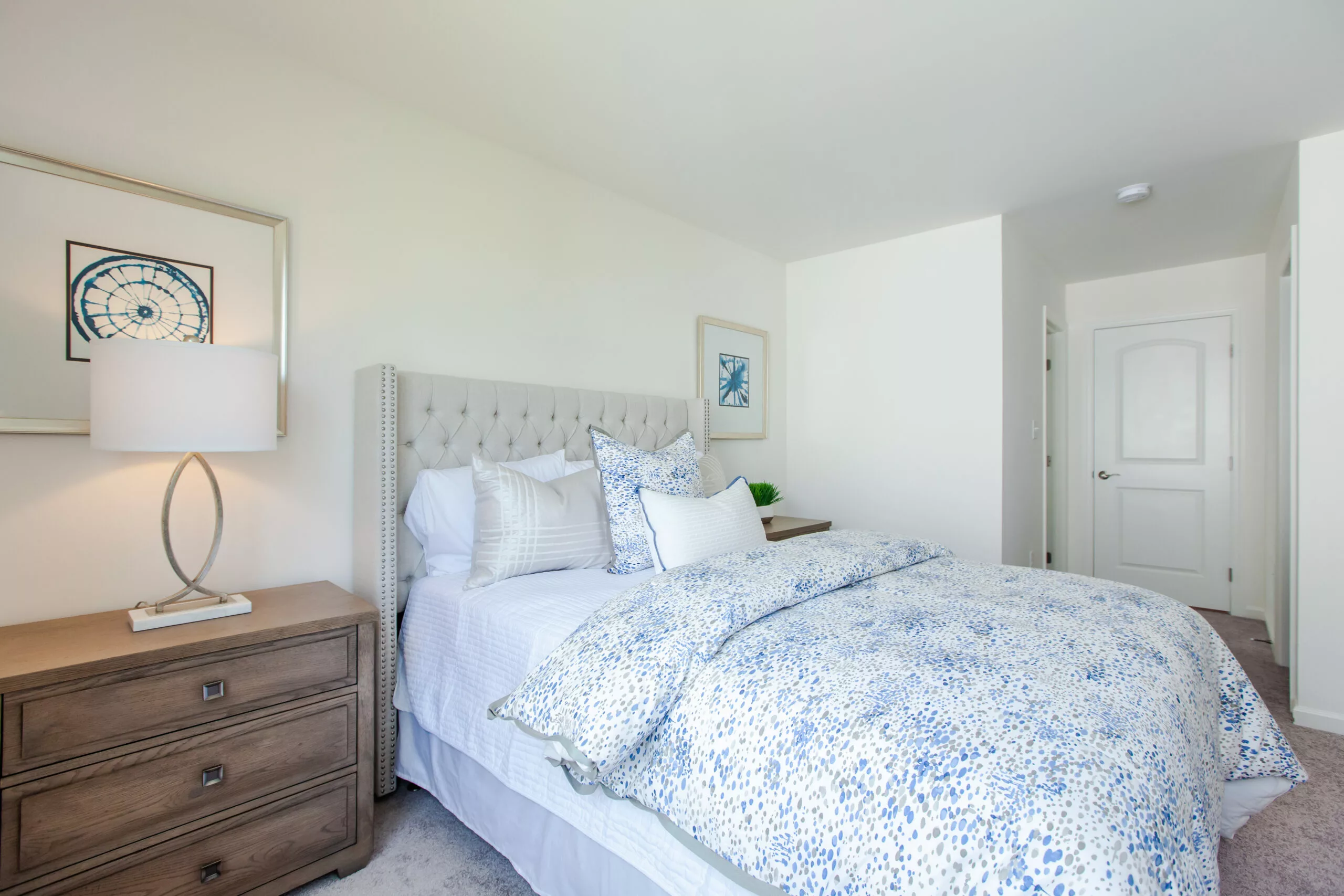 A bright bedroom with white walls, a tufted beige headboard, blue and white patterned bedding, wooden nightstand with a lamp, and framed artwork. An open doorway leads to a hallway.