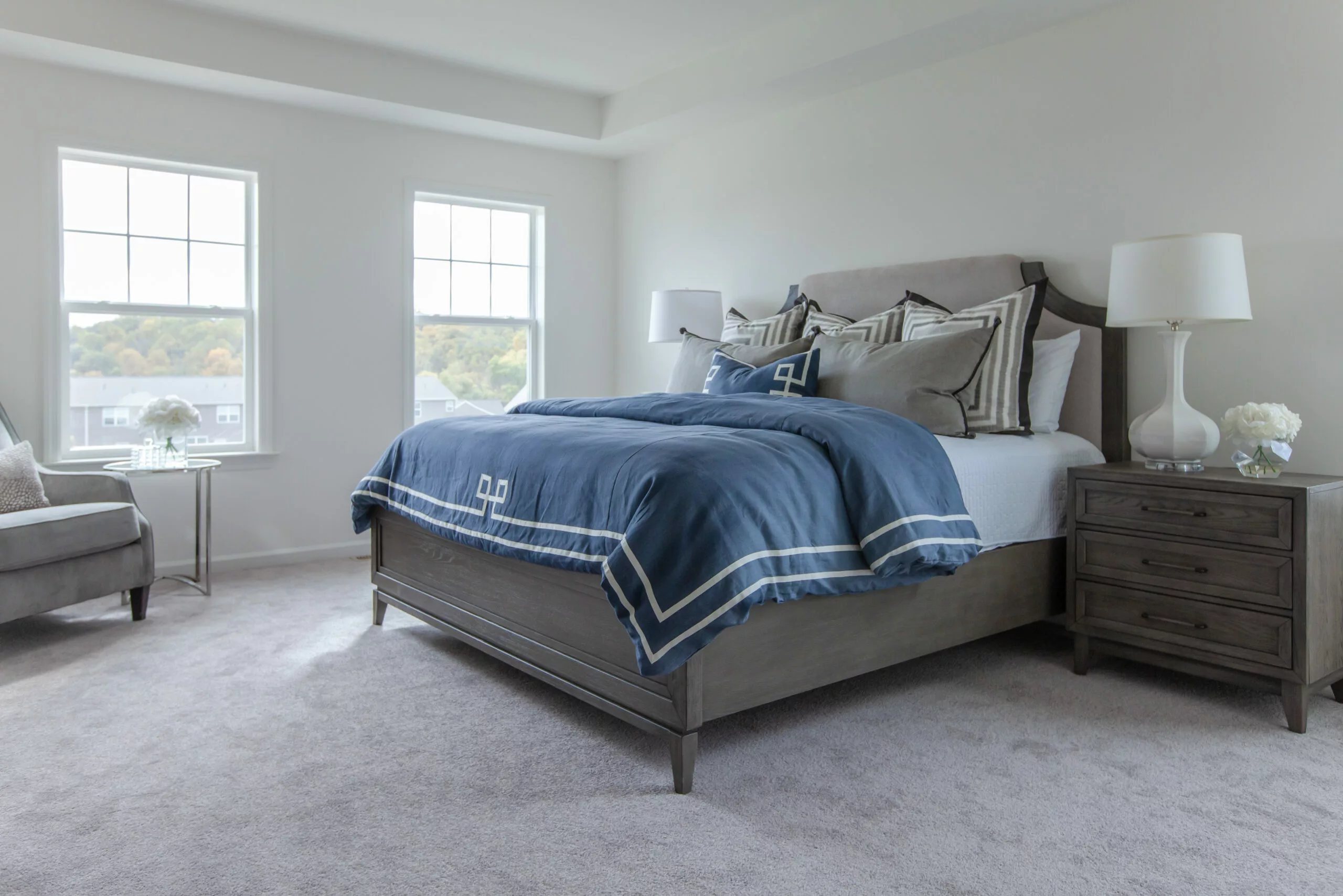 A bright, modern bedroom with large windows, a gray bed frame, blue and white bedding, matching nightstands with white lamps, and a gray armchair on light carpet flooring.