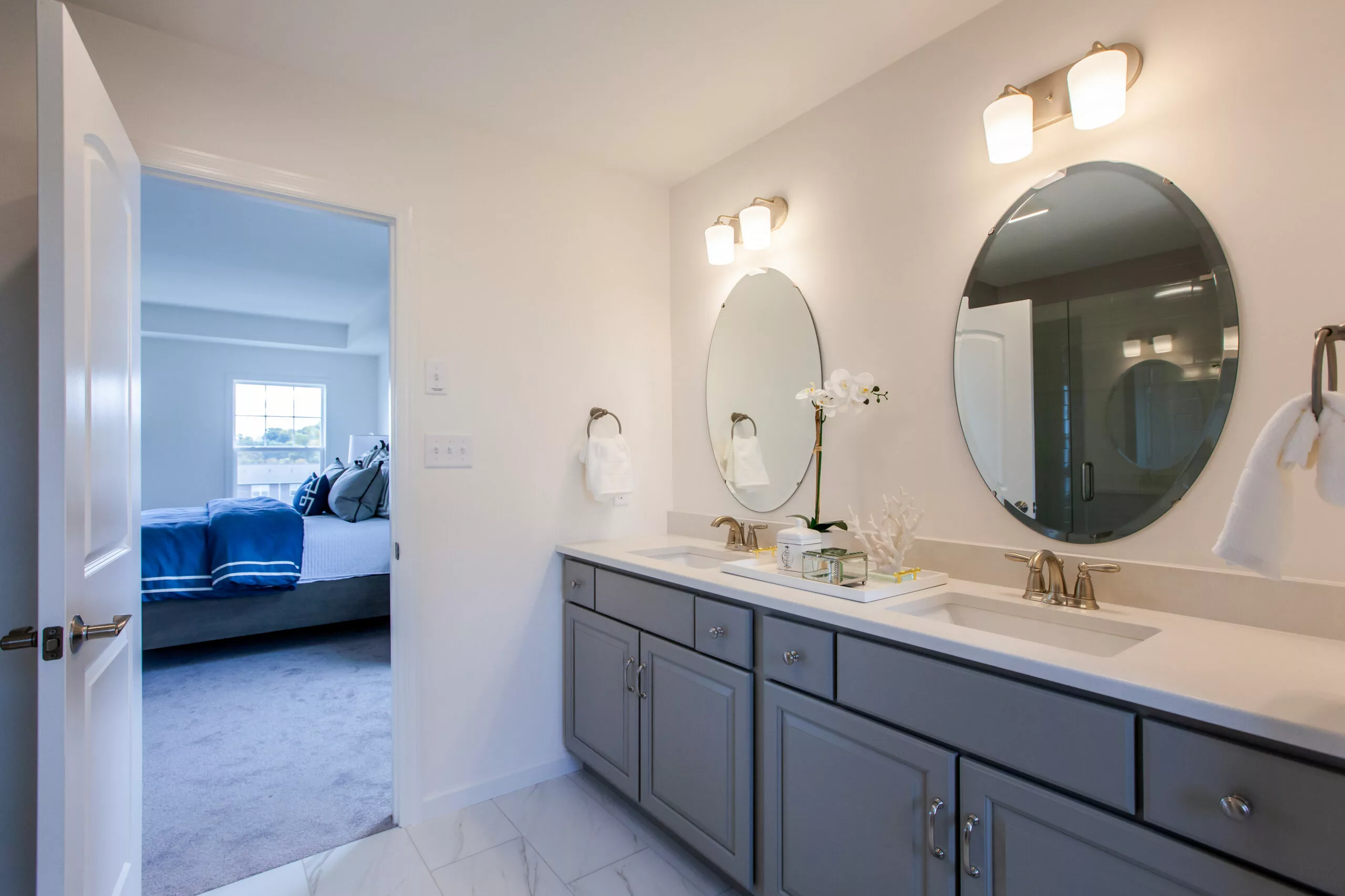A modern bathroom with double sinks, gray cabinets, oval mirrors, and wall lights. The counters have decorative items and towels. An open door leads to a bedroom with a bed and window in the background.