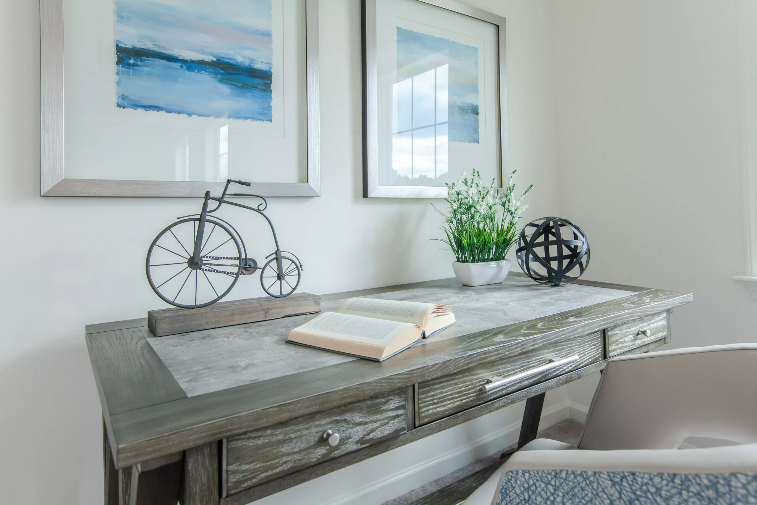 A modern wooden desk with an open book, a potted plant, a decorative bicycle, and a black sphere. Two abstract blue and white paintings hang on the wall above the desk. A white chair is partially visible.