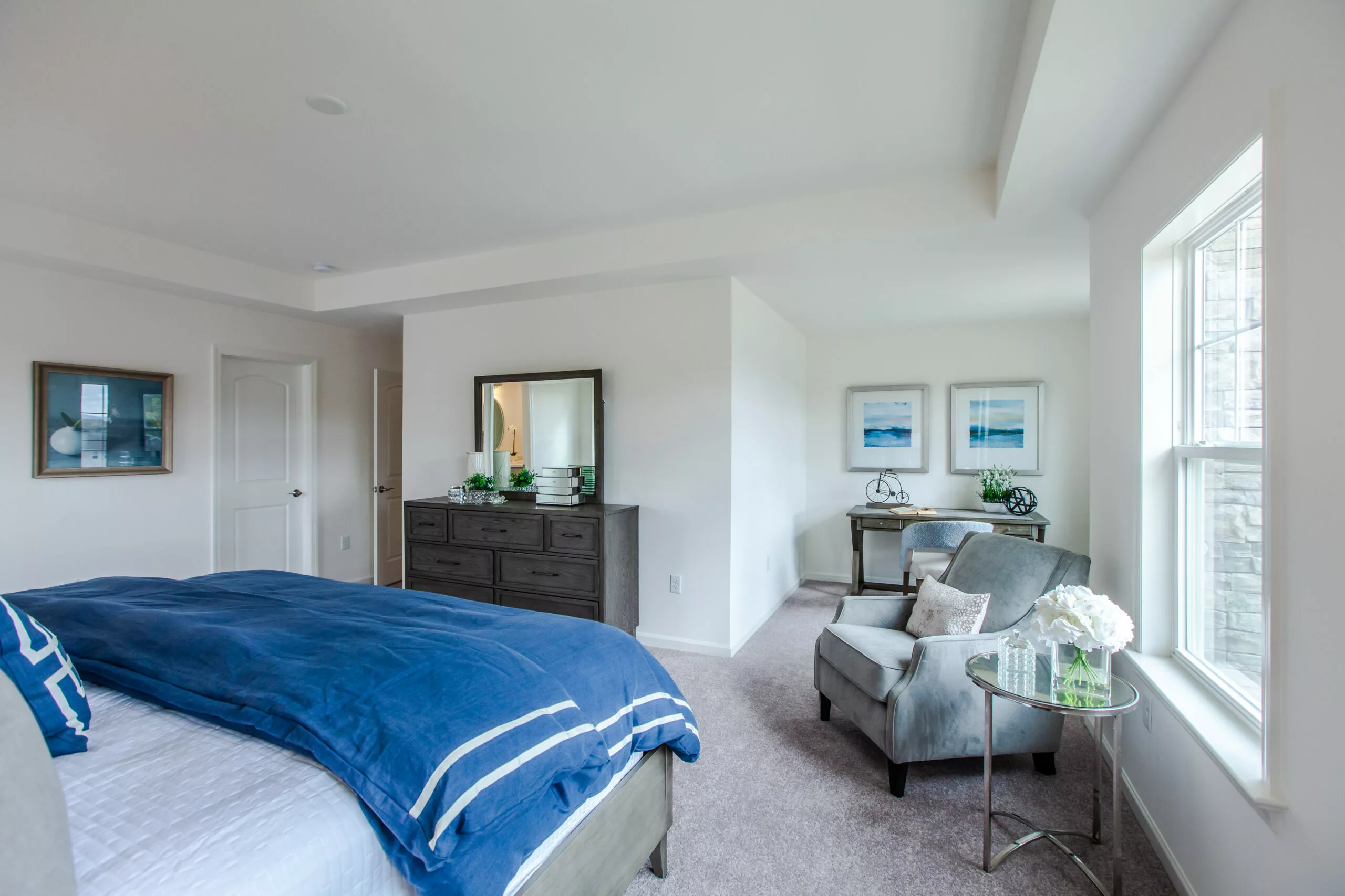 A bright, spacious bedroom with a large bed covered in blue and white bedding, a dark dresser with a mirror, a small sitting area with two armchairs, a side table with flowers, and large windows letting in natural light.