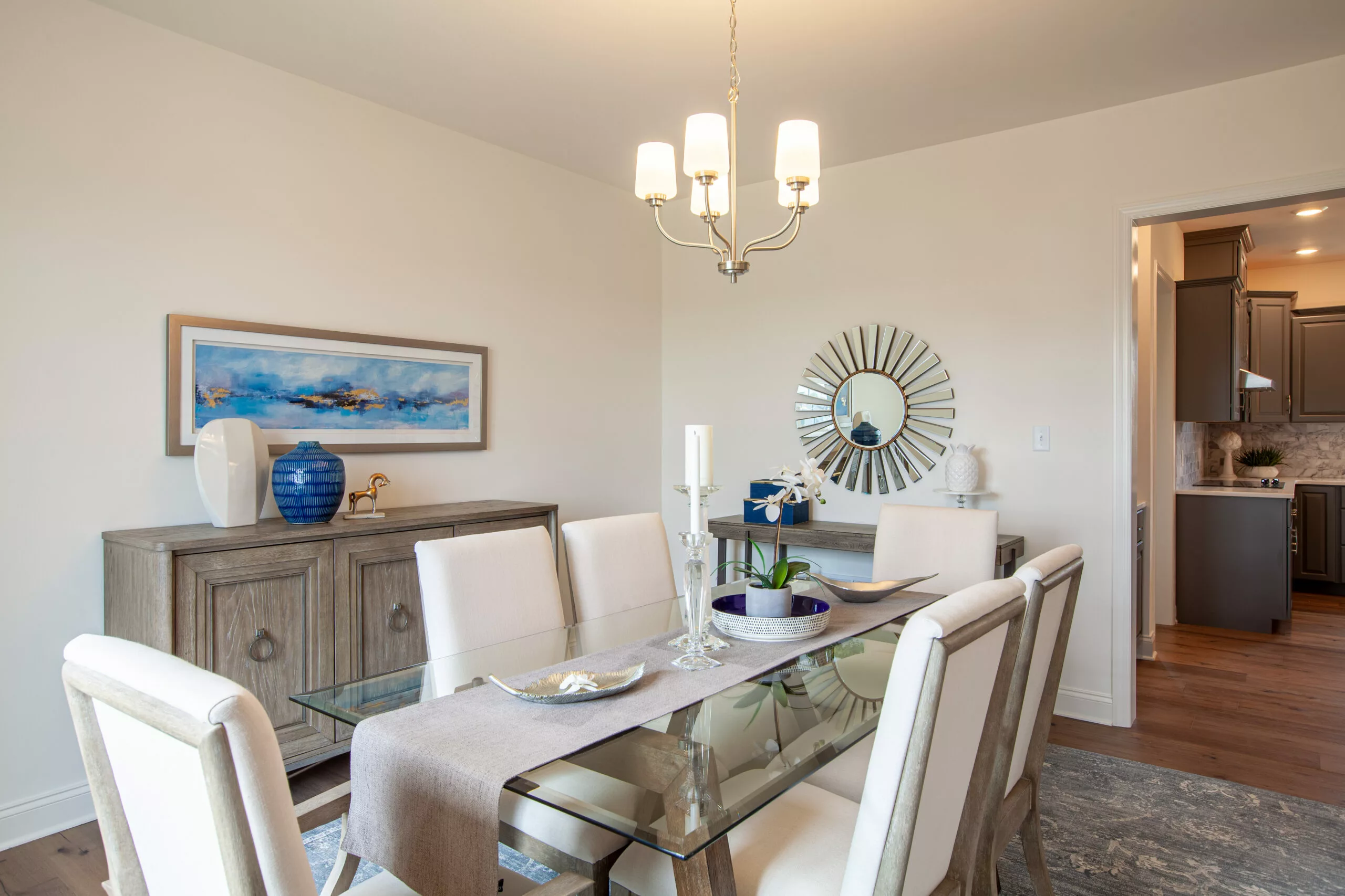 A modern dining room with a glass table, six white chairs, a light fixture above, and a sideboard decorated with a blue vase, candles, and artwork. A sunburst mirror hangs on the wall near a doorway to the kitchen.